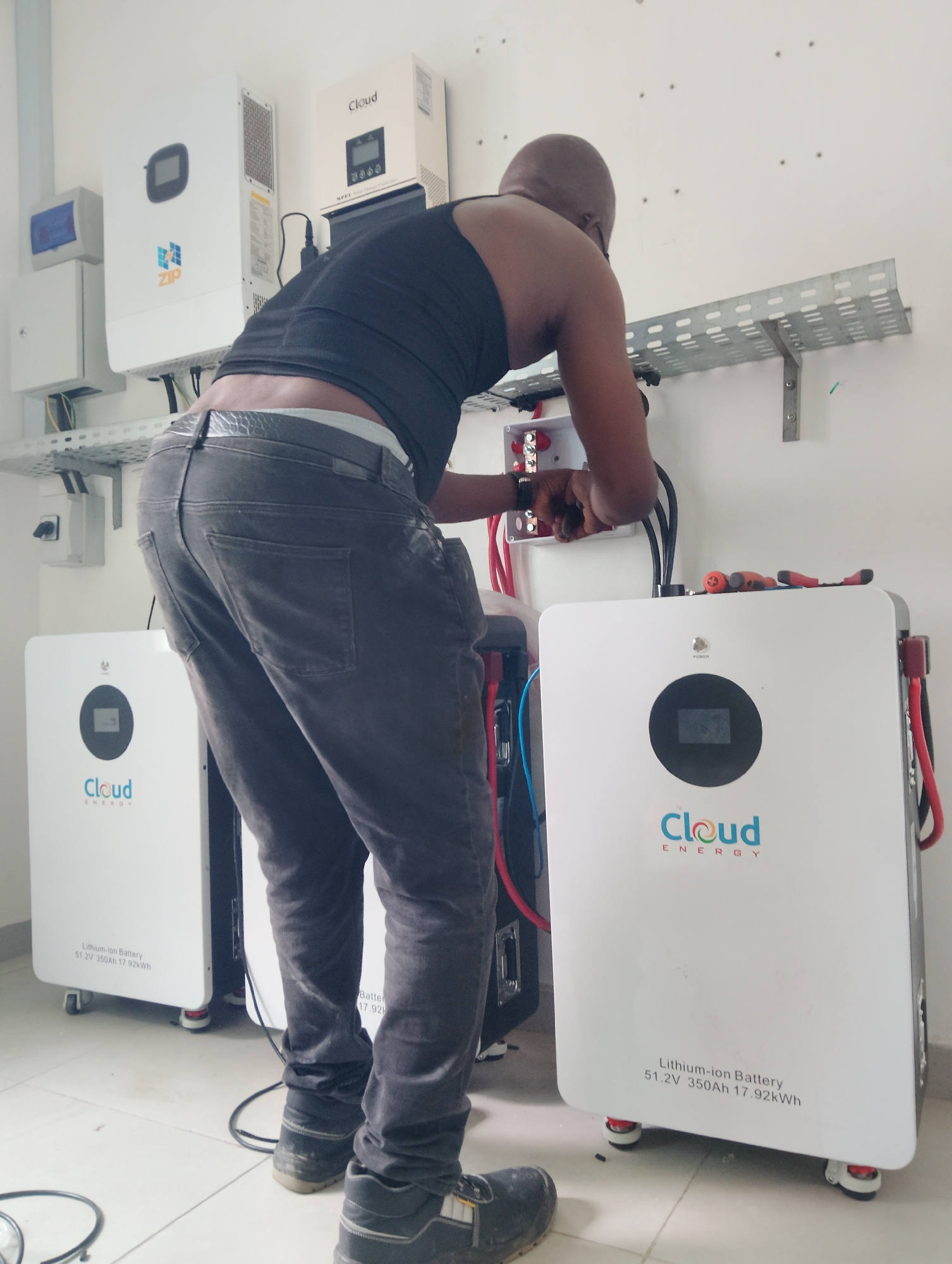 In a room with white tiled floors and white walls, a man is working on a solar power system. He is wearing a black tank top and dark grey jeans. The man is bent over, connecting wires to a white battery unit labeled "Cloud ENERGY". There are two such battery units visible in the image, along with a white solar inverter labeled "ZIP" and another smaller white unit. A metal cable tray is mounted on the wall above the equipment. The scene appears to be in a commercial or residential building in Ikorodu, Nigeria, possibly an electrical installation site or a storage area for solar equipment.