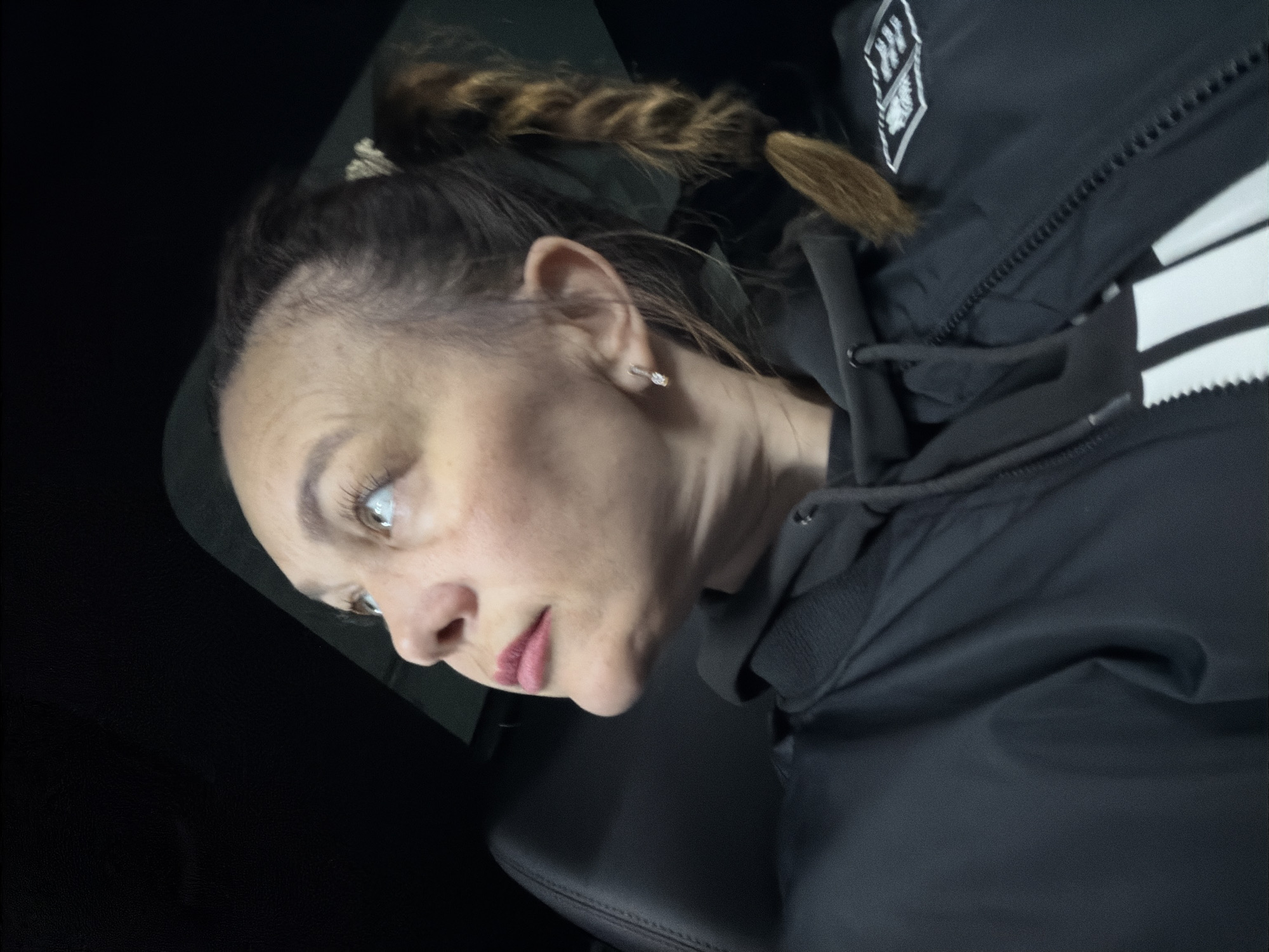 A medium close-up portrait shows a woman in a dimly lit environment, likely inside a vehicle, as suggested by the visible black car seat and interior. The image is taken from a slightly tilted angle.
The woman has fair skin, light-colored eyes, and is wearing light pink lipstick. Her hair, a mix of dark and light brown/auburn, is pulled back, with a prominent braid visible over her right shoulder (viewer's left). A small, sparkling stud earring is visible in her left ear. She has a neutral expression, gazing slightly off-camera to her left. Some fine lines are visible on her forehead.
She is dressed in a black hooded jacket or coat. The jacket features a white emblem or logo on the upper left chest, which clearly displays the letters "N" and "W," along with white stripes on the right sleeve. The fabric of the jacket appears to be a practical, possibly water-resistant material.
The background is dark and indistinct, reinforcing the impression of an interior space. The overall lighting is dim, suggesting it could be evening, night, or an enclosed space. The scene depicts a casual moment, potentially while waiting or traveling in Chaikovsky, Russia, as per the provided context.
