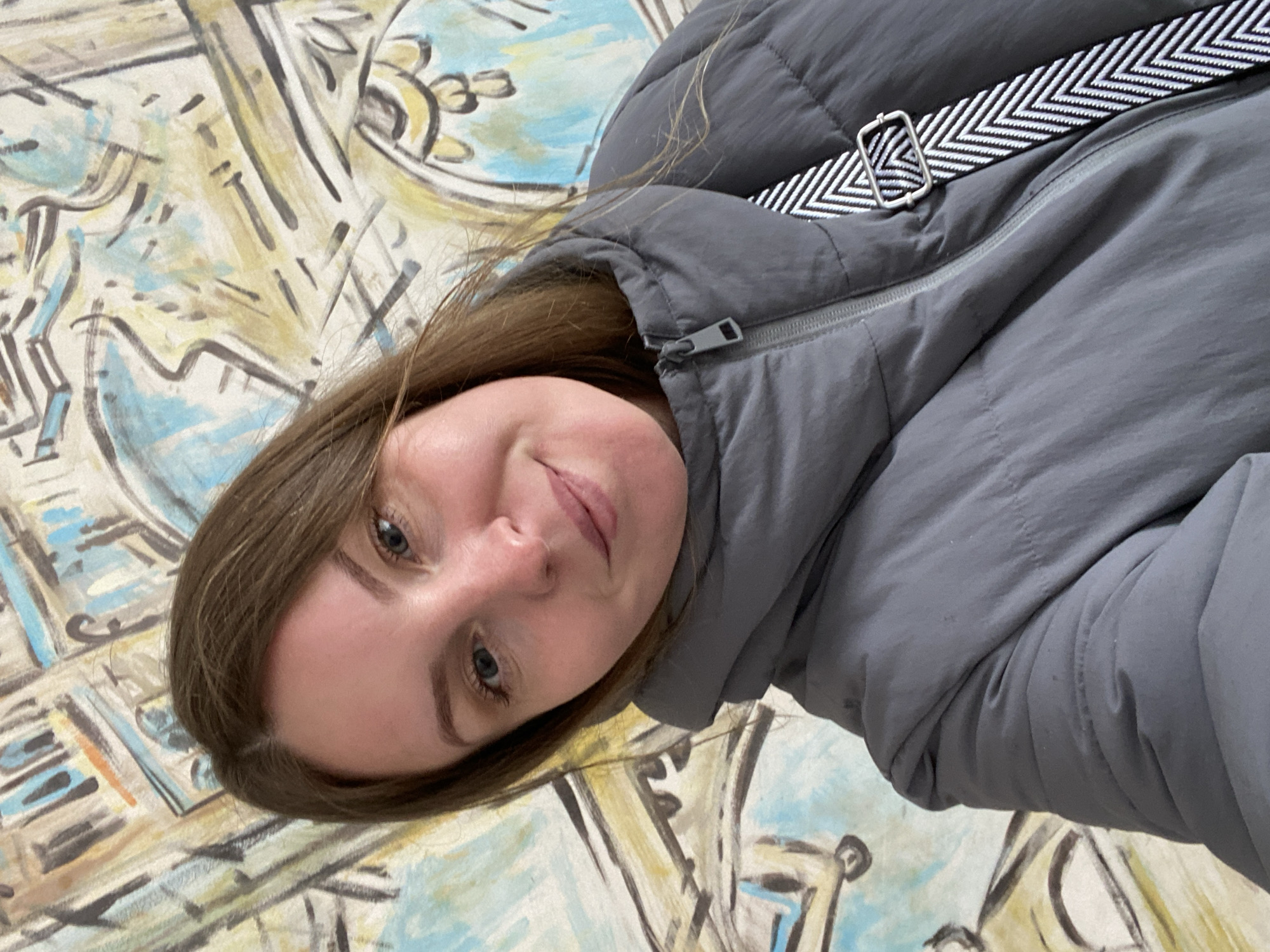 A young woman is captured in a close-up portrait, with the image rotated 90 degrees counter-clockwise. She has light brown hair, blue eyes, and fair skin, and exhibits a gentle, closed-lip smile, conveying a calm and pleasant demeanor. She is wearing a grey padded or puffer jacket, which features a visible zipper and a silver-toned zipper pull at her neck. A distinct black and white zigzag-patterned strap is visible over her right shoulder (as if the image were correctly oriented), secured by a rectangular, silver-colored buckle.
The background consists of a colorful, abstract mural or painting covering a wall. This artwork is characterized by stylized lines and shapes, outlined in dark brown or black, and filled with washes of light blue, beige, and yellow. The artistic style suggests an indoor setting, possibly a gallery, a public space, or a decorated room in Chaikovsky, Russia. The lighting is even, further indicating an interior environment. There is no visible text in the image.