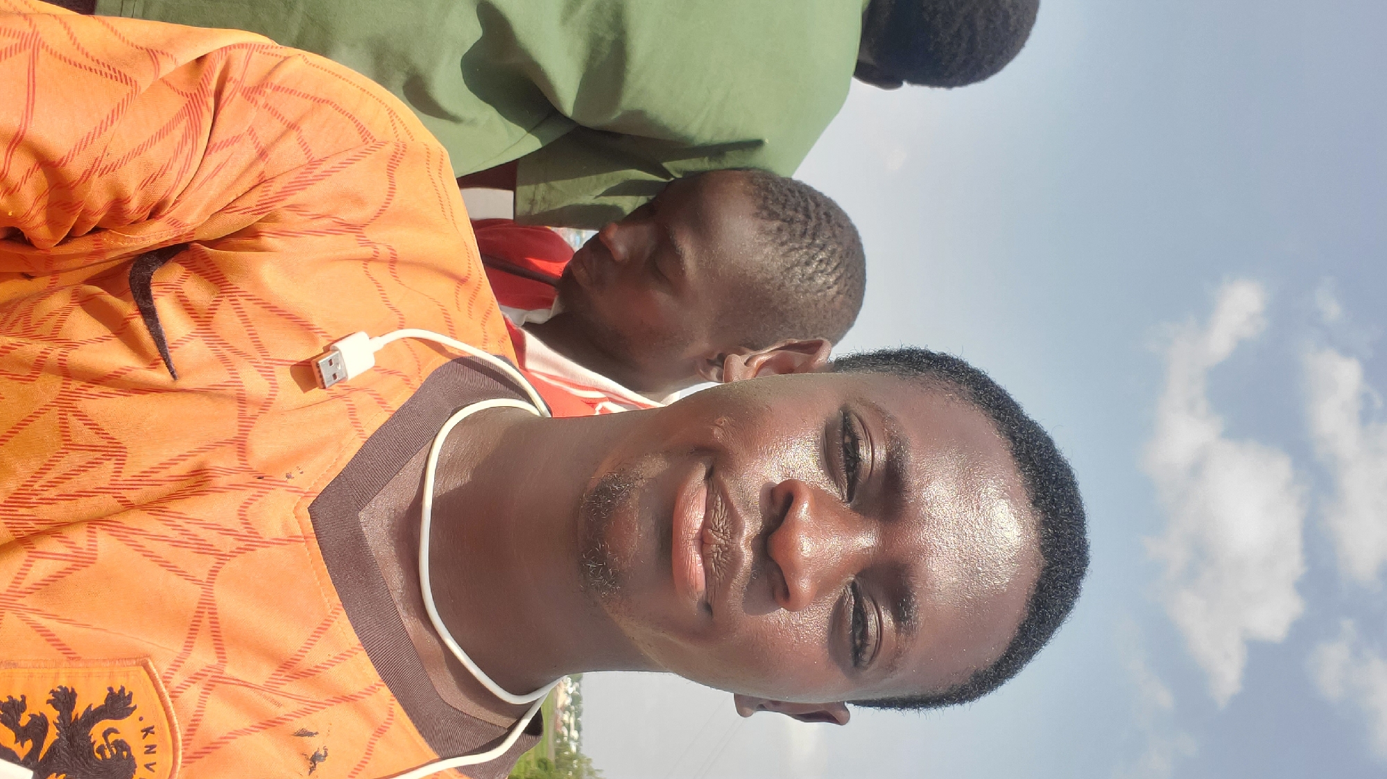 In Jalingo, Nigeria, a young man is captured in a selfie, his face glistening with sweat under the bright sun. He wears an orange jersey with a black Nike logo and a Dutch KNVB crest. A white cord, connected to a USB-like device, loops around his neck and rests on his jersey. Behind him, two other young men are visible. One, in a green shirt, is partially obscured, while the other, wearing a red and white striped shirt, looks down, his face partially shadowed. The background suggests an outdoor urban setting with a bright blue sky dotted with white clouds.