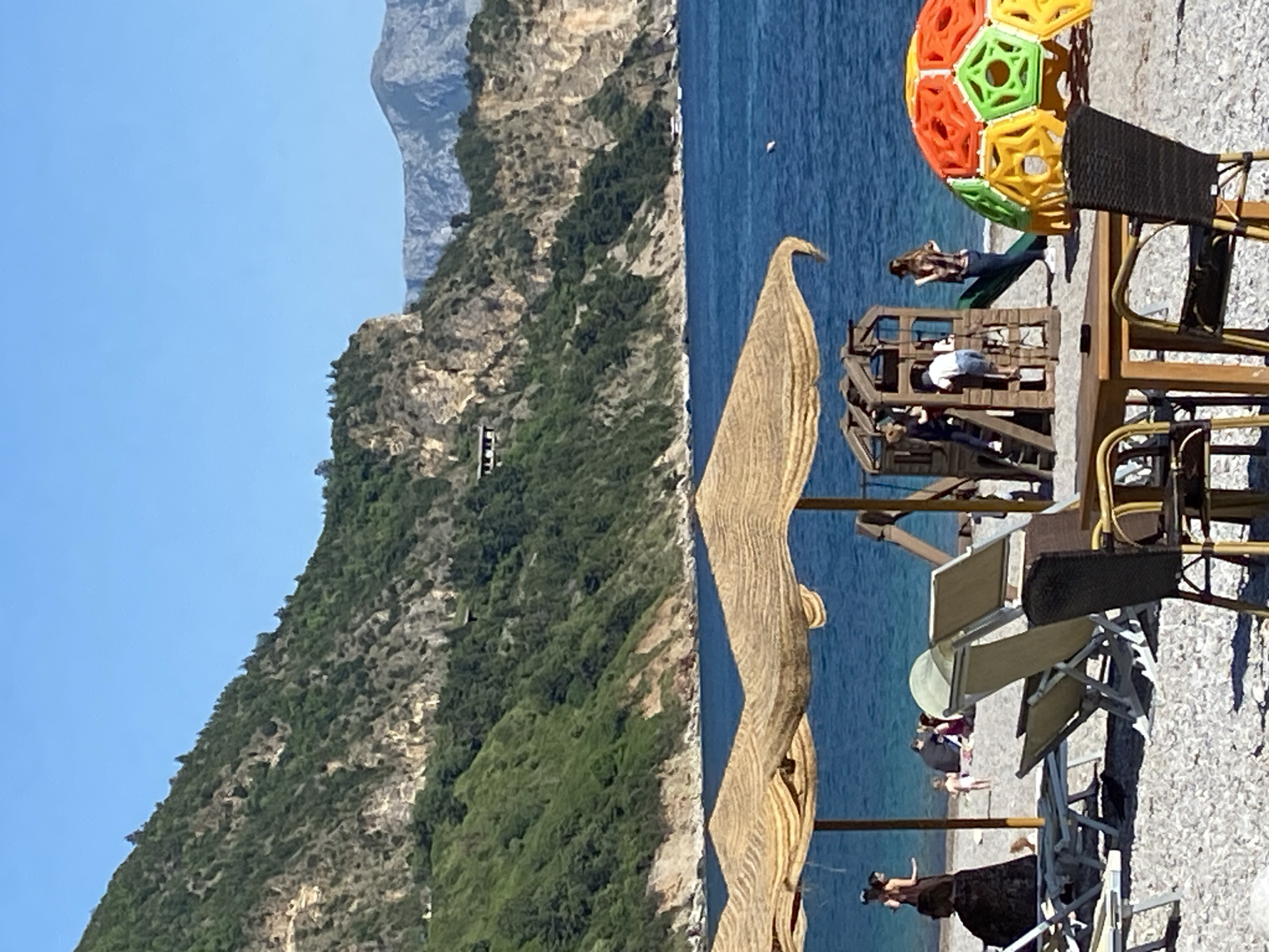 The image depicts a coastal scene in Prijevor, Montenegro. The foreground showcases beachside amenities, including lounge chairs, tables, and a distinctive wicker structure resembling a heron. A multi-colored, geometric playground structure with people on it is visible, along with other beachgoers scattered along the shore. The background features a rugged, mountainous landscape covered in lush green vegetation, rising steeply from the coastline, and a clear blue sky above. The sea, a deep blue color, contrasts sharply with the light-colored beach.