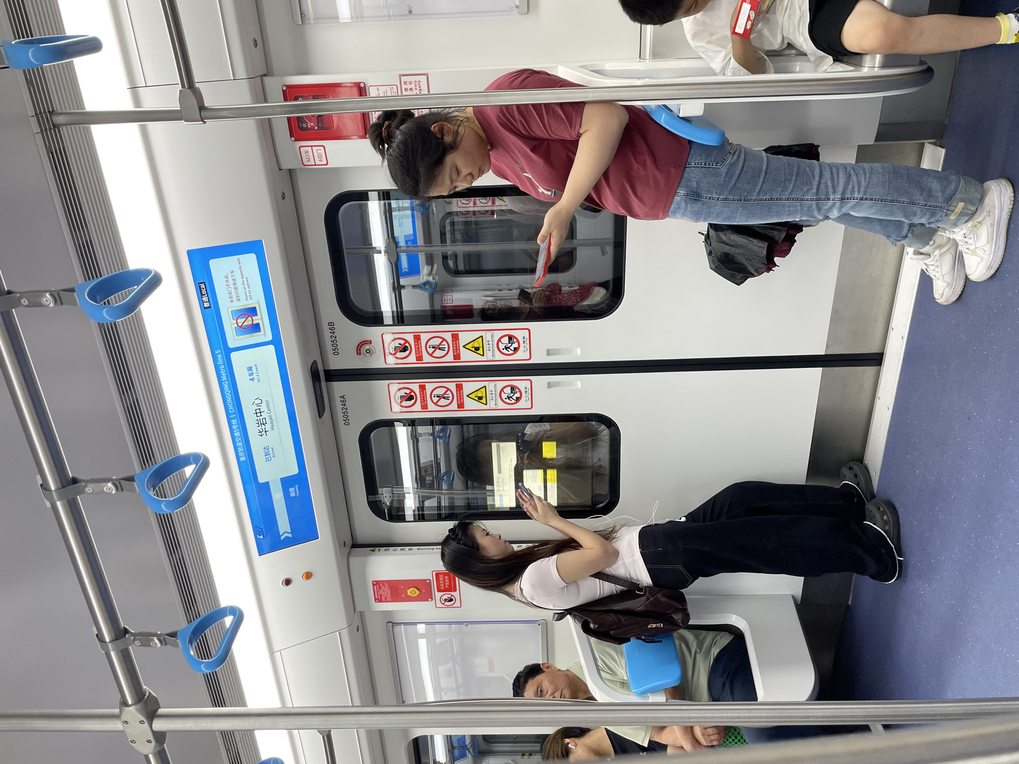The image shows the interior of a subway car. A woman in a red shirt and blue jeans stands holding a red phone. Next to her, a child is seated. The woman appears to be occupied with the device. Another woman, wearing a white t-shirt and black pants, stands facing the door, also using her phone. Another person sits below her and to her left. Overhead, blue grab handles extend downward from the ceiling. A transit advertisement is posted on the door, and some text on the advertisement references Chongqing Metro Line 5 and mentions "Huayan Center", which is a station in the Jiulongpo District of China. The door is white. Various safety icons are displayed on the door above the window. A label indicates "M2车" and "6位门."