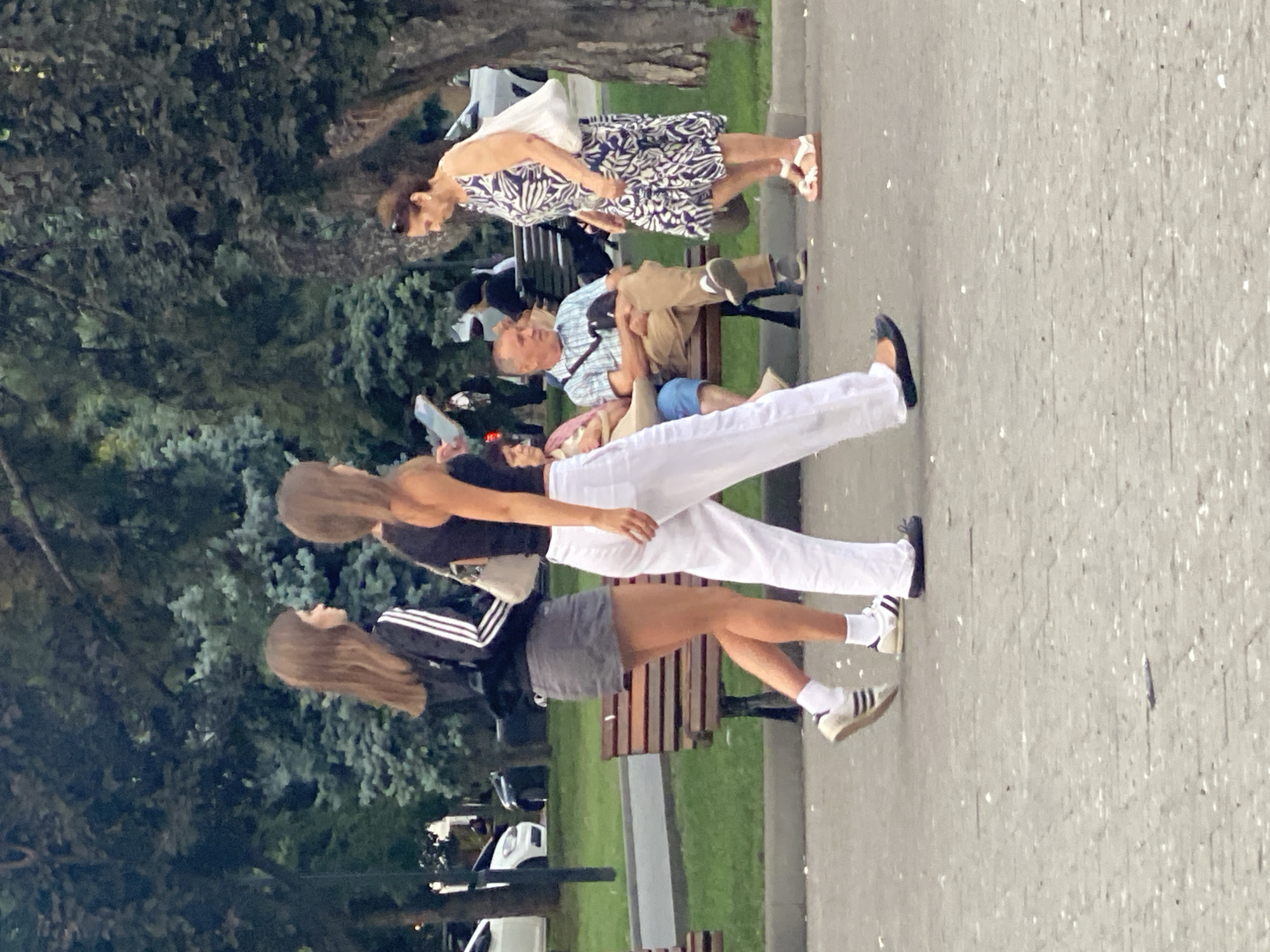The image depicts an outdoor scene in a park or public square, likely in Chișinău, Moldova. In the foreground, two young women are walking side-by-side on a paved pathway. The woman on the left wears a black athletic jacket with white stripes, a grey skirt, white socks, and white and black sneakers. She has long brown hair. The woman on the right wears a black tank top and loose white pants, with black flat shoes. She also has long brown hair.
In the middle ground, a wooden park bench is occupied by three individuals. A man in a checkered shirt and light brown pants is seated, with a dark bag slung across his chest. Next to him, a woman with dark hair is partially visible, wearing a light pink top. To the right of the man, another woman is standing. She is wearing a patterned blue and white dress and white sandals. She is looking down.
In the background, lush green trees form a dense canopy. Several cars are visible through the trees, suggesting the proximity of a road or parking area. The overall impression is a casual gathering in a public green space.