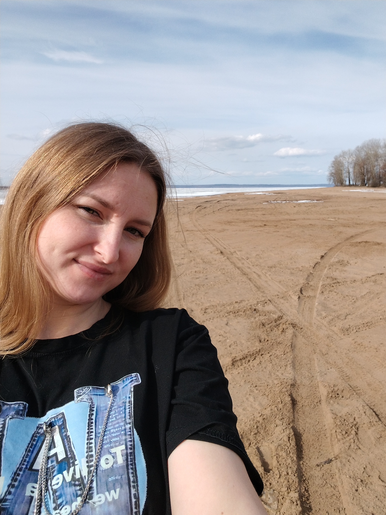 A woman is taking a selfie outdoors. She has light brown hair and is wearing a black t-shirt. Behind her is a wide, sandy beach. In the distance, there is water with patches of ice and a line of trees. There are tire tracks in the sand. The sky is partly cloudy. The location appears to be a beach in Noviy, Russia.