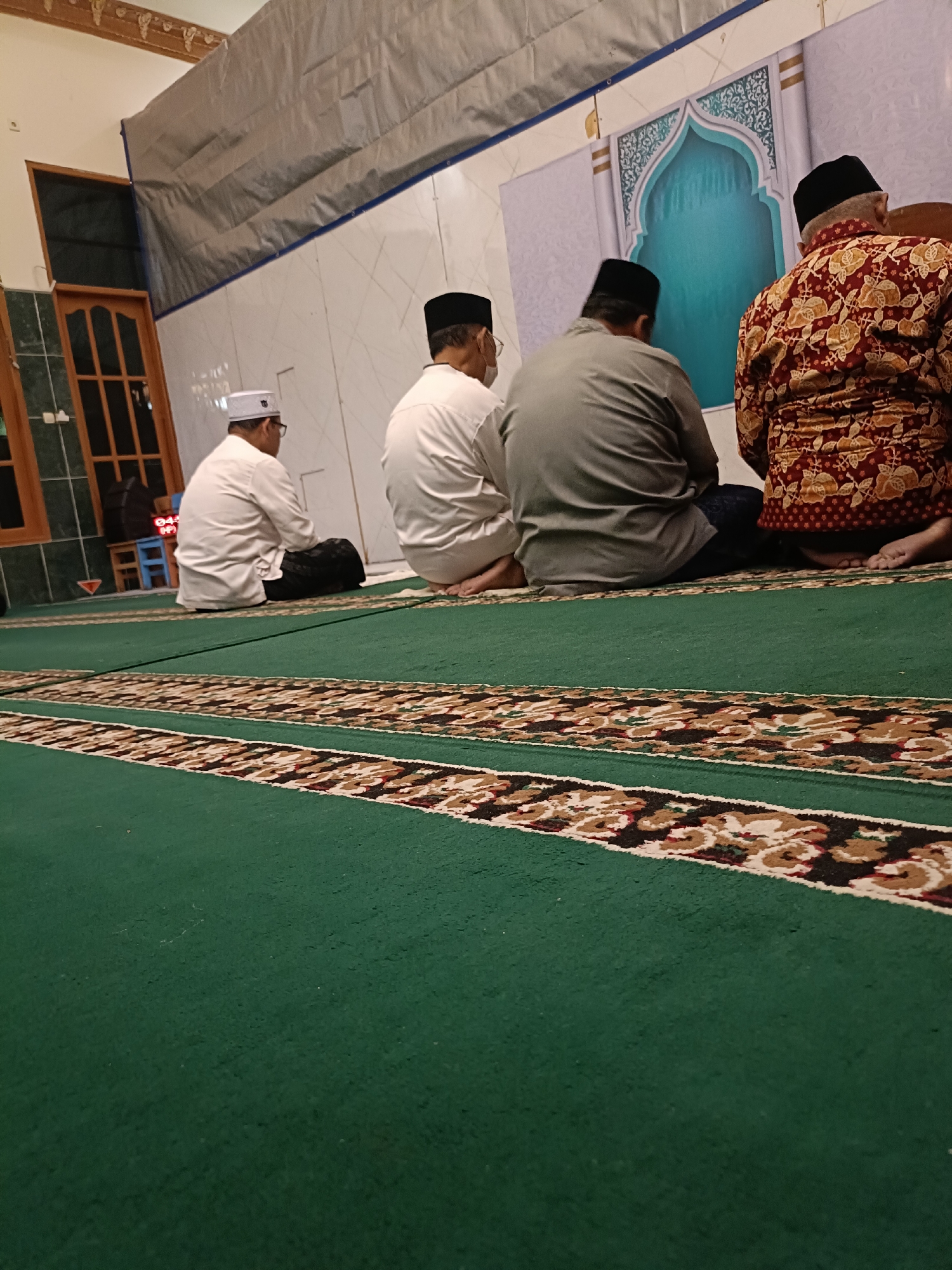 Four men are seated on a green carpet in what appears to be a mosque or prayer room. The men are facing away from the camera, towards a decorative wall that resembles an arched doorway. Three of the men are wearing white caps, and one is wearing a black cap. The man on the far right is wearing a patterned maroon and gold shirt. The man second from the left is wearing a grey shirt, and the two men to his left are wearing white shirts. The floor is covered in a green carpet with a decorative border. In the background, there are windows and a draped sheet. The image appears to have been taken at night or in low light conditions, likely in Kediri City, Indonesia.