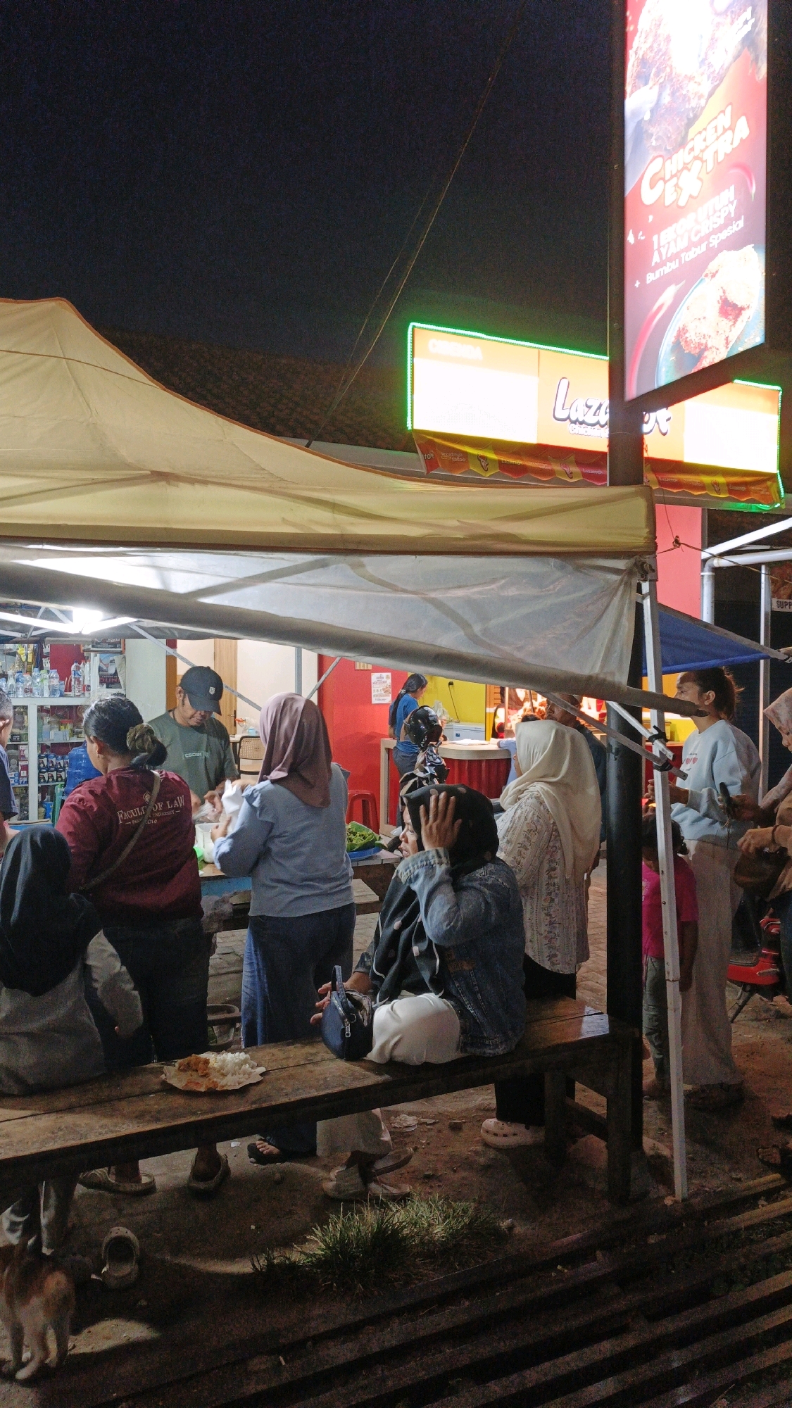 This image depicts an outdoor scene at night, likely in Cibenda, Indonesia, with a focus on a food stall or eatery. A group of people are gathered under a makeshift canopy, illuminated by artificial light sources, including bright neon signs. One prominent vertical sign displays text and images related to chicken, with "CHICKEN EXTRA" being clearly visible. Another horizontal sign with green neon outlining reads "Lazus."
Several individuals are visible, some interacting with a stall or counter, while others are seated on a wooden bench. A plate of food, appearing to be rice and some other items, is placed on the bench. People are dressed in casual attire, with some wearing headscarves, suggesting a cultural context. The environment appears to be an informal setting, possibly a street food area or a local market, with some debris and uneven ground visible. In the lower left corner, a cat is partially visible. The overall impression is a lively night scene centered around food consumption and social interaction in a local Indonesian setting.