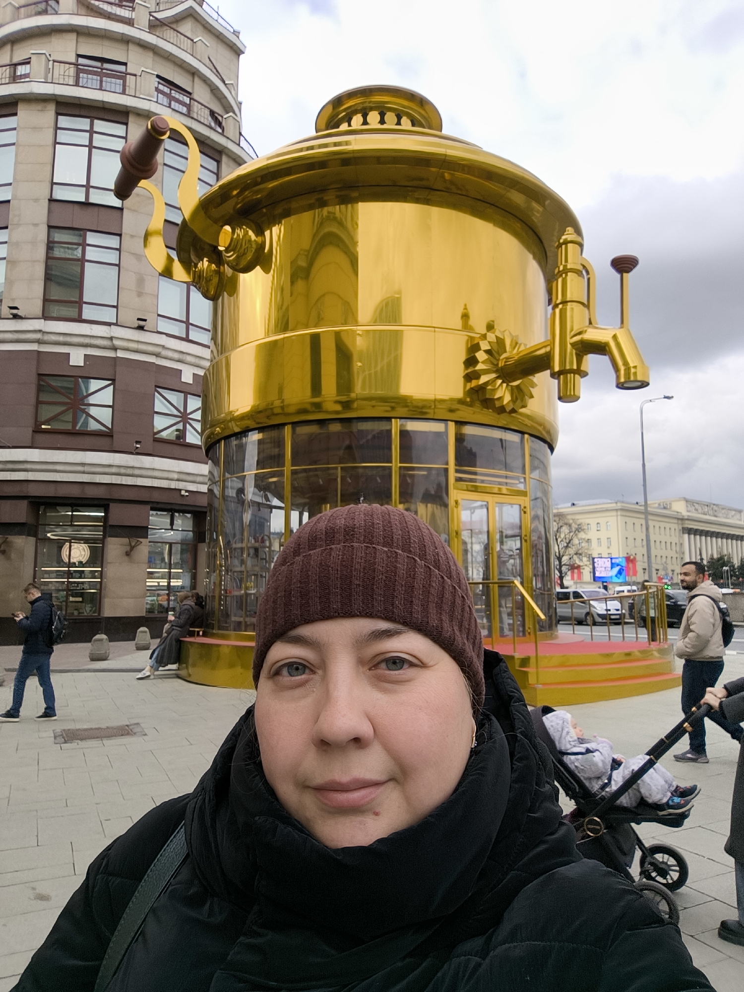 This urban selfie captures a woman in the foreground against a backdrop dominated by a colossal golden samovar-shaped building in Moscow, Russia. The woman, positioned on the left, wears a brown beanie and a black puffer jacket, looking at the camera with a neutral expression.
The central feature is the multi-story, highly polished golden structure resembling a traditional Russian samovar, complete with an ornate handle on the left, a spout, and a prominent tap on the right. Its base is a glass-enclosed section, suggesting it functions as a cafe or commercial space, known locally as the "Tula Samovar" cafe on Tverskaya Street.
In the background, to the left of the golden samovar, stands a tall, rounded building with numerous windows and balconies. Further right, in the distance, a large classical-style building with columns is visible. The scene includes several other people: a man walking on the left, two individuals sitting near the samovar, a man with a backpack on the right, and a child in a stroller being pushed by an unseen person.
The setting is a paved pedestrian area adjacent to a city street, under an overcast sky, suggesting a cool daytime. No discernible text is visible.