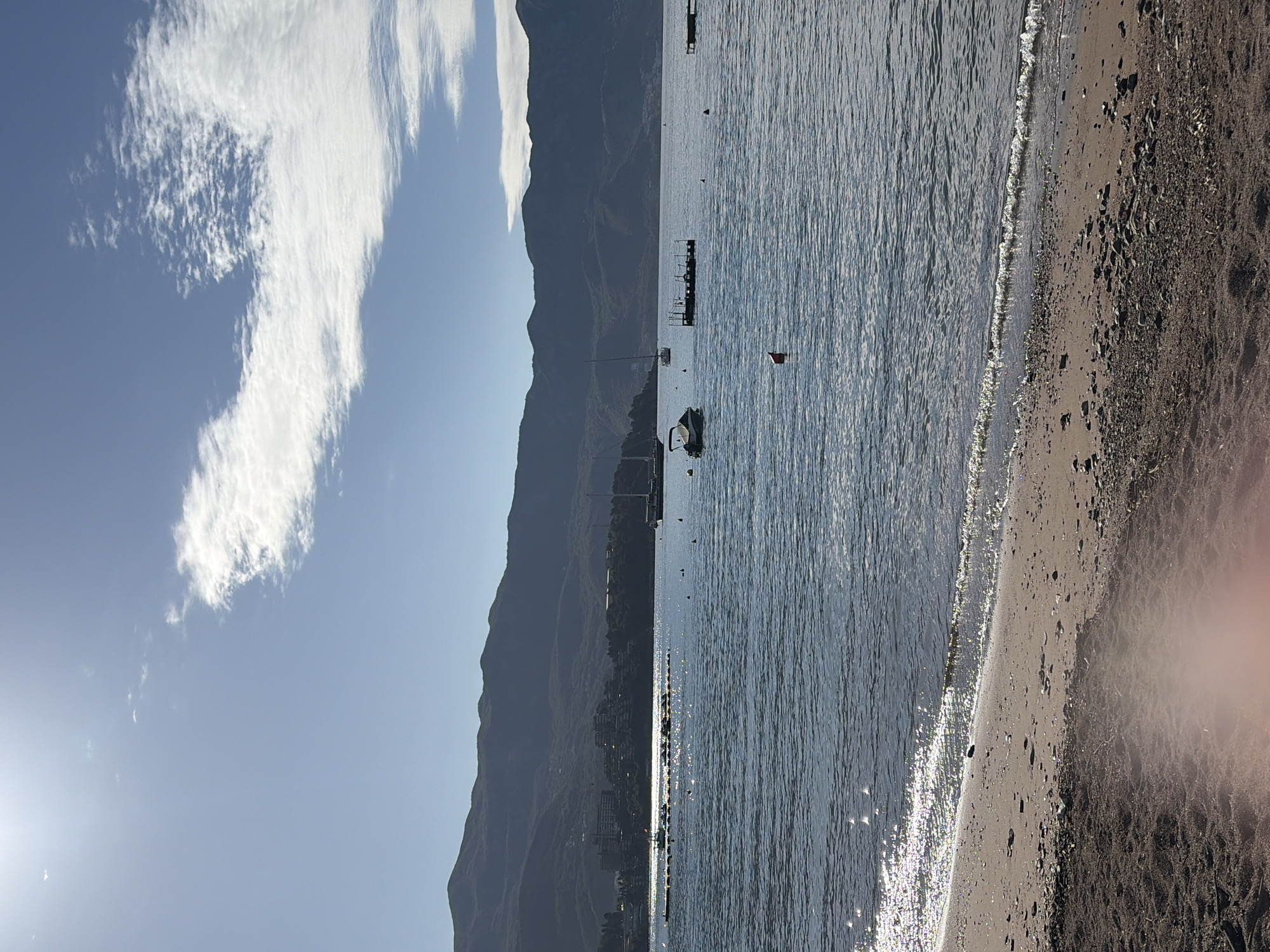 The image depicts a coastal scene in Budva, Montenegro, oriented vertically. In the foreground, a beach with dark sand transitions to a lighter, wet sand area near the water's edge. The water is a dark blue with shimmering sunlight reflecting off its surface. Further out in the water, there are boats and structures, including a small pier and a buoy. The background features a mountain range that stretches along the coastline, partially obscured by haze. The sky is light blue with wispy white clouds extending vertically upward. The sun is visible as a bright glare in the lower left corner.