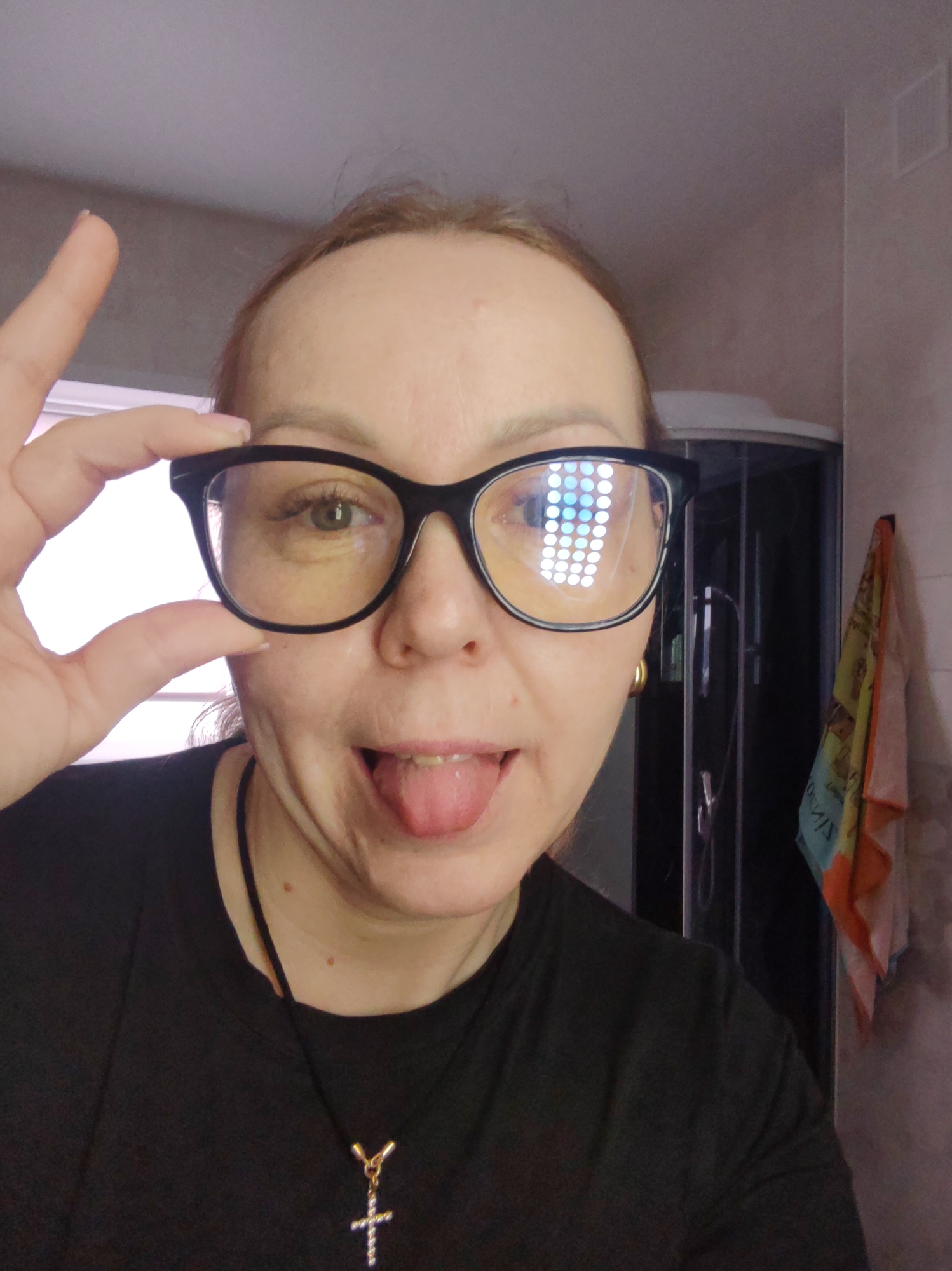 A Caucasian woman with light-brown hair pulled back, wearing black-rimmed glasses, a black t-shirt, and a gold cross necklace, is taking a selfie in what appears to be a bathroom. She is sticking her tongue out and making a pinching gesture with her fingers around the frame of her glasses. The reflection in her glasses suggests she is near a source of bright, square lighting. In the background is a shower stall and a patterned towel. The location is not discernible from the image.