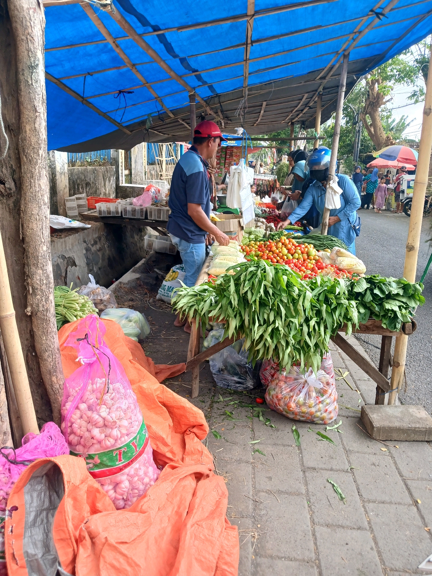 This is a photograph of a street-side produce stall in Banyuwangi, Indonesia. A vendor, a man wearing a red cap and blue shirt, is serving customers. He is standing behind a wooden table laden with fresh vegetables, including tomatoes, green beans, corn, and leafy greens. Two customers, a woman in a blue head covering and mask, and another person in a blue head covering, are interacting with the vendor, appearing to select produce. In the foreground, large bags of garlic and other produce are visible, some wrapped in orange tarpaulin. The stall is covered by a blue tarp, supported by bamboo poles. The background shows a street with a few more people and stalls, suggesting a local market or busy street corner. The ground is paved with concrete tiles, and some debris and fallen leaves are scattered around the stall. The scene depicts a typical day at a local market in a bustling urban environment.