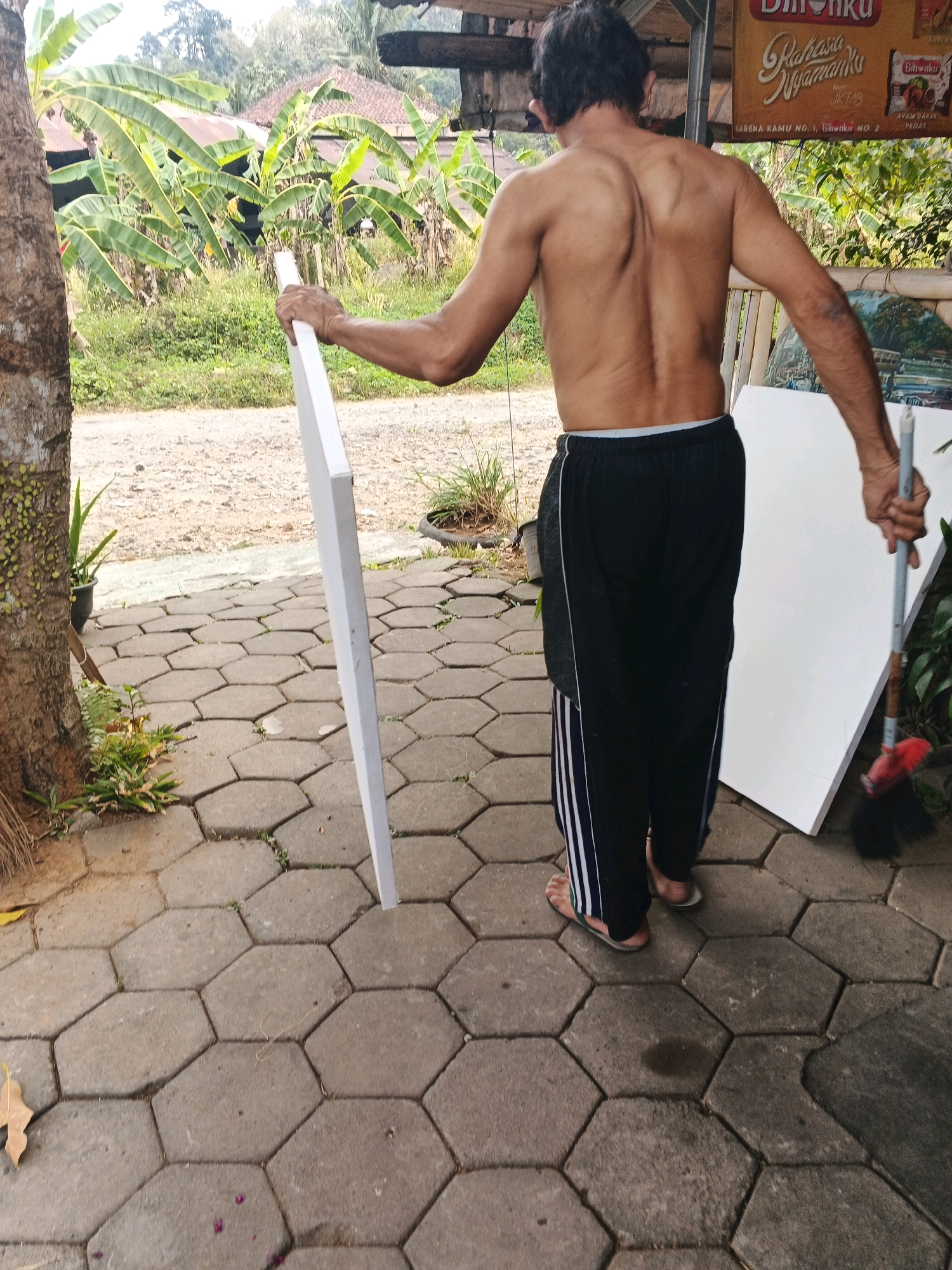 A bare-chested man, facing away from the camera, stands on a hexagonal paver patio. He is holding a large, flat, white object vertically in his left hand, its lower end resting on the pavers. His right hand is holding a broom with a red brush head, angled downwards towards the ground. He is wearing black athletic pants with white stripes down the sides and flip-flops. Behind him, banana trees and a thatched-roof structure are visible, suggesting a semi-rural or outdoor area. In the background, a sign with Indonesian text and images of food products is partially visible. The overall scene appears to be set in a residential or commercial outdoor space in Ungaran, Indonesia.