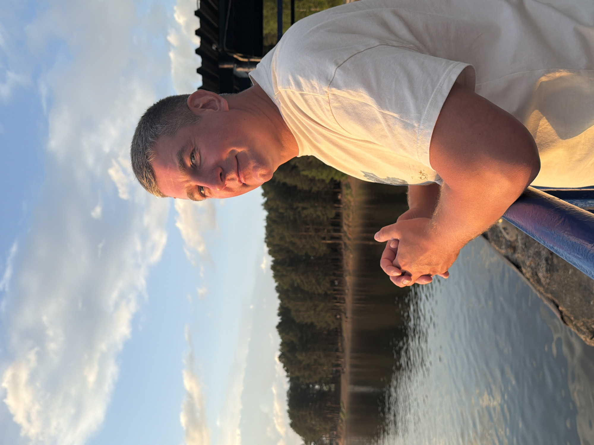 A middle-aged man is photographed outdoors during the evening. He has short, graying hair and wears a white T-shirt. He leans on a blue railing, looking toward the camera with a slight smile. Behind him, there is a view of a lake with trees lining the shore. The sky is blue with scattered clouds. Given the context, the location is possibly the city of Chaikovsky, Russia.