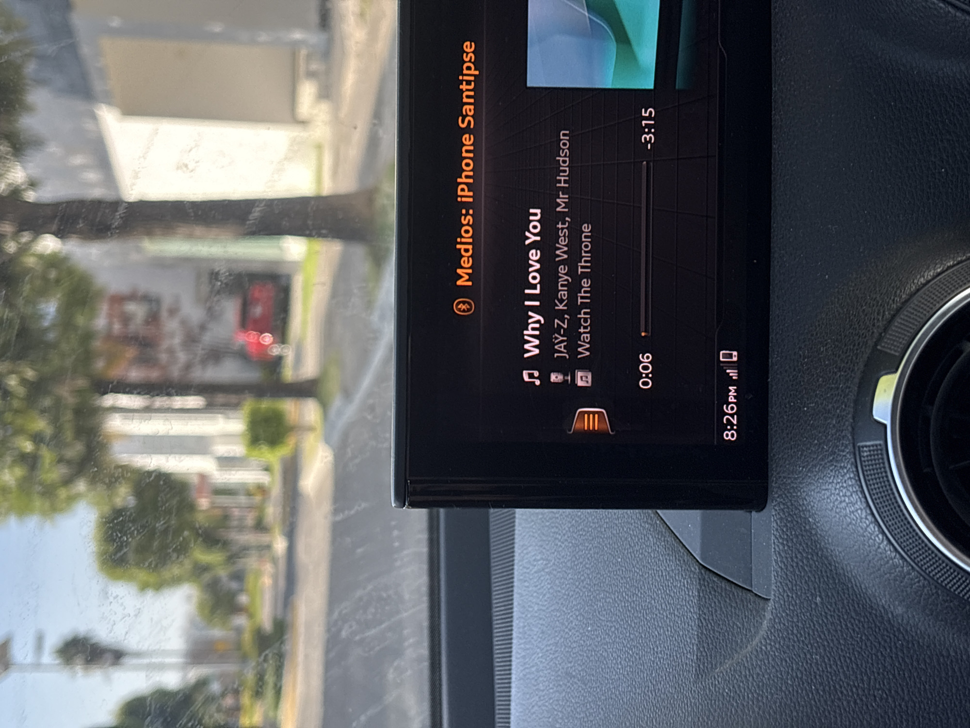 The image captures a close-up view of a car's infotainment system displaying music playback information. The screen shows the title "Why I Love You" with the artists JAY-Z, Kanye West, and Mr. Hudson, and the album "Watch The Throne." The song progress is at 0:06, with a total duration of -3:15. The system is connected to "Medios: iPhone Santipse." The time displayed on the car's dashboard is 8:26 PM. In the background, seen through the windshield, is a blurred street scene with trees, buildings, and a red car. The environment appears to be outdoors during daylight. The car's interior shows a textured dashboard and a circular vent. The overall impression is a snapshot of a car journey in an urban or suburban setting. The reference to San Bernardino Tlaxcalancingo, Mexico, is not visually present in the image itself but is provided as a context for the location.