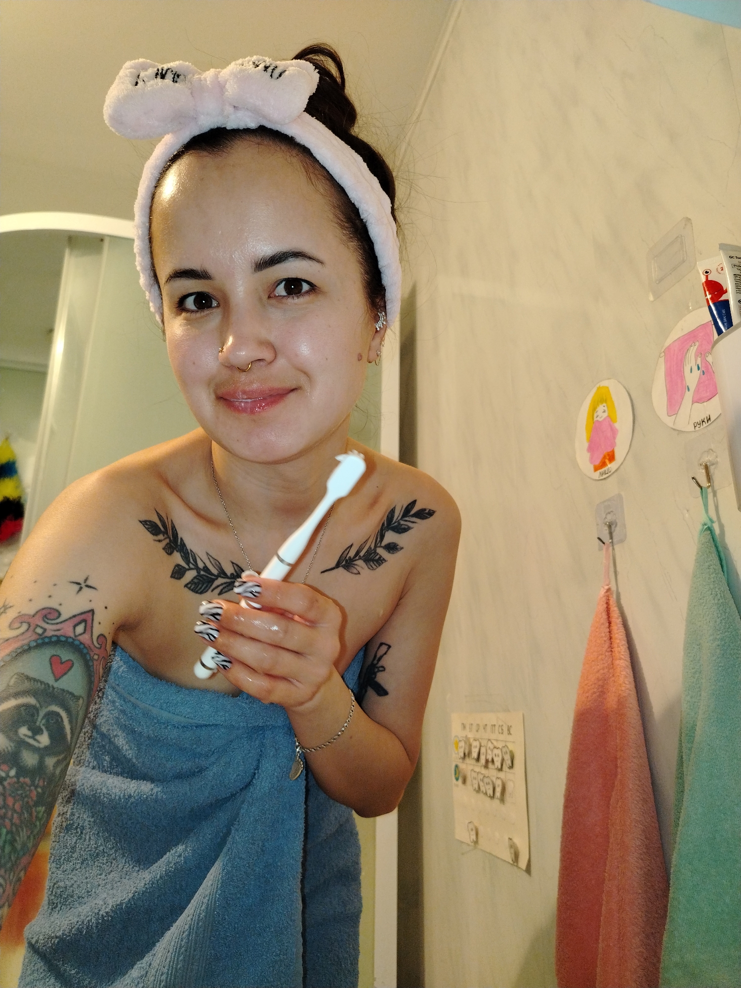 A woman with dark hair, wearing a pink fuzzy bow headband, is captured in a close-up, waist-up portrait in what appears to be a bathroom setting. She is wrapped in a blue towel and holds a white electric toothbrush in her right hand. Her fingernails are painted with a black and white zebra print. She has a septum piercing, a nostril stud, and multiple ear piercings, and smiles gently at the camera. Visible tattoos include leafy branches across her chest and a colorful, detailed design featuring a racoon and a heart on her left arm.
The background shows a light-colored wall with various bathroom items. To the right, two small hand-drawn circular decorations are affixed to the wall; one depicts a yellow figure, the other a hand washing with bubbles, labeled with the Cyrillic word "РУКИ" (hands). Below these are two small, colorful towels (one orange/pink, one teal/light green) hanging on hooks. A clear plastic holder is attached to the wall, revealing part of a tube labeled "GC Tooth M" and "Professional." Further right, a white shower enclosure is partially visible. Another piece of paper with drawings of teeth/faces, possibly a dental hygiene chart, is visible with the Cyrillic text "ПН ВТ СР ЧТ ПТ СБ ВС" (Monday, Tuesday, Wednesday, Thursday, Friday, Saturday, Sunday). The woman's pink headband also has partially visible text that reads "BABY GIRL." The overall impression is a personal moment of self-care.