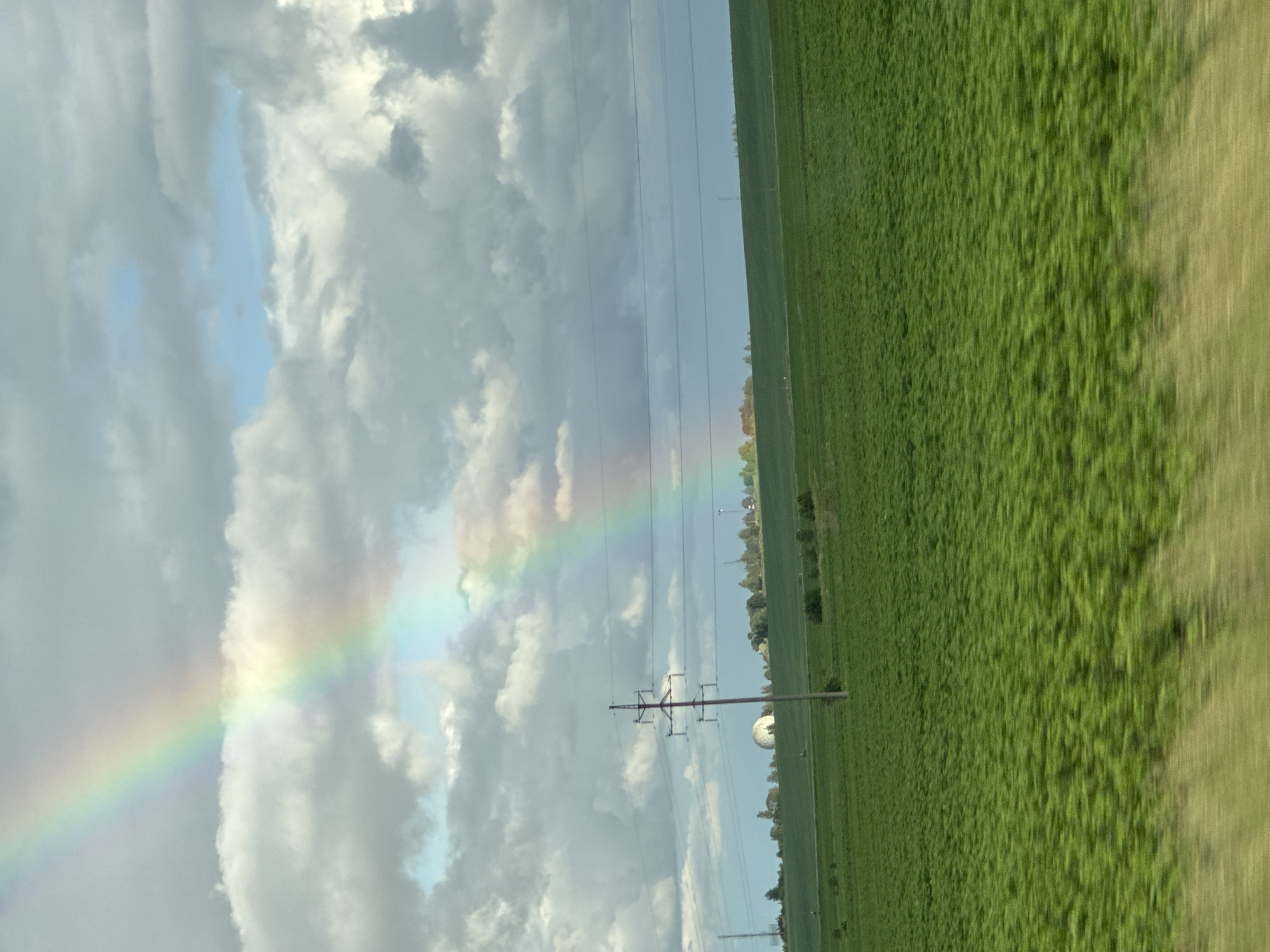 The image depicts a rural landscape under a cloudy sky with a prominent rainbow arching across the upper portion. The foreground is dominated by a vast, green field, likely agricultural, with a sense of motion blur suggesting the photograph was taken from a moving vehicle. In the mid-ground, a series of power lines are strung between utility poles, one of which is a prominent feature on the left. Beyond the field and power lines, a gentle slope rises, dotted with trees and a distinctive white, domed structure, possibly a radar station or a scientific facility. The sky is a mix of grey and white clouds, with patches of blue sky visible, and the rainbow displays a spectrum of colors. The overall impression is one of a scenic, possibly post-rainy day in a countryside setting. The specific location within Borskoe, Russia, is not visually indicated in the image itself.