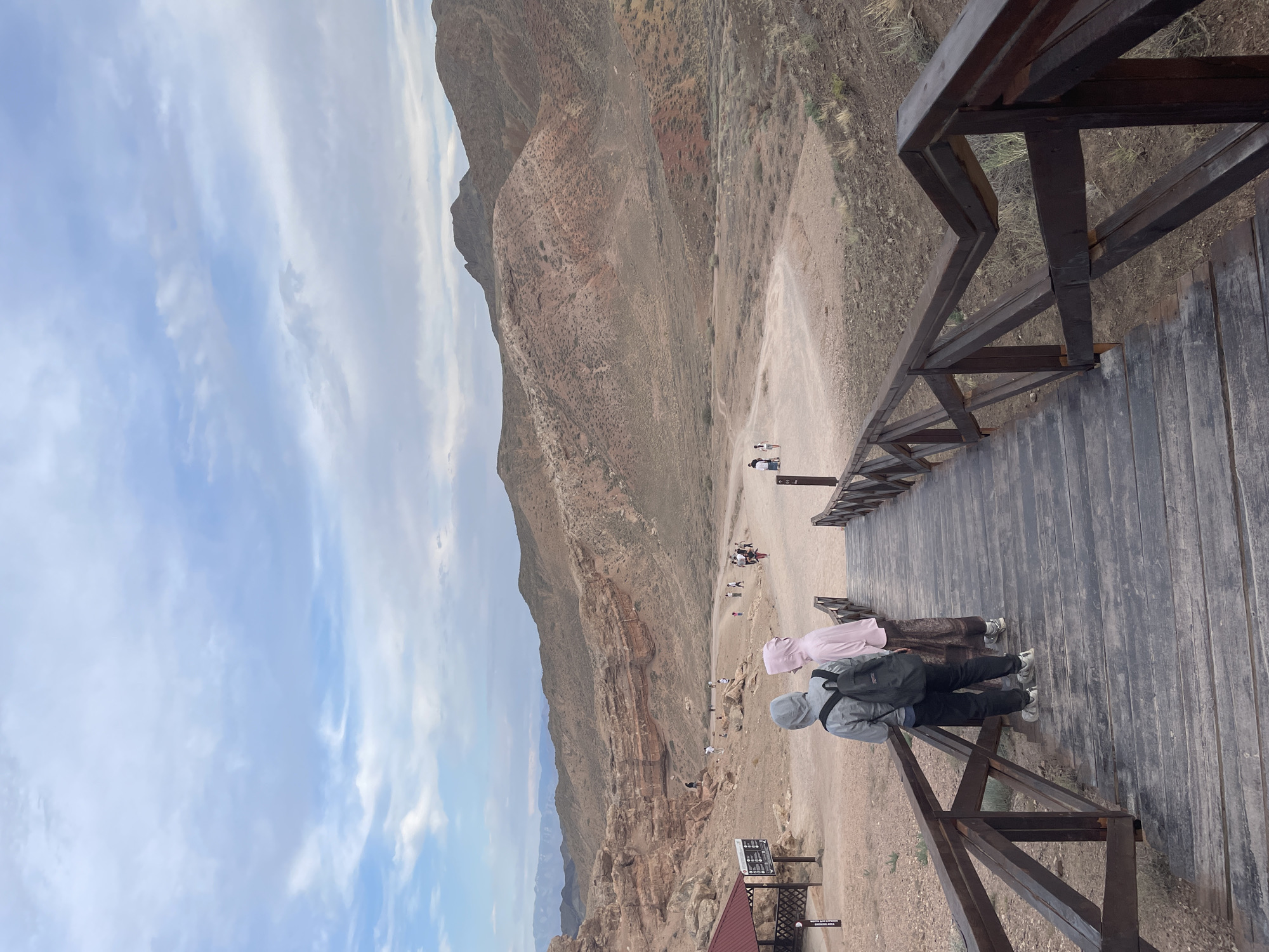 The image depicts a scenic view of the Charyn Canyon, a natural park located in the Almaty Region of Kazakhstan, near the city of Tamerlik. In the foreground, a wooden staircase with a railing leads down into the canyon. Two individuals, one wearing a grey hooded jacket and a black backpack, and another in a pink hooded jacket and a patterned skirt, are standing on the stairs, facing away from the camera. Further down the path, several other individuals are visible walking and exploring the canyon. The canyon itself is characterized by its steep, eroded walls of reddish-brown rock formations, with a dusty trail winding through the valley. The sky above is partly cloudy, with a mix of blue and white. A small red-roofed structure, possibly a visitor information point or shelter, is situated on the left side of the frame near the top of the stairs. The overall impression is a natural landscape with signs of human visitation.