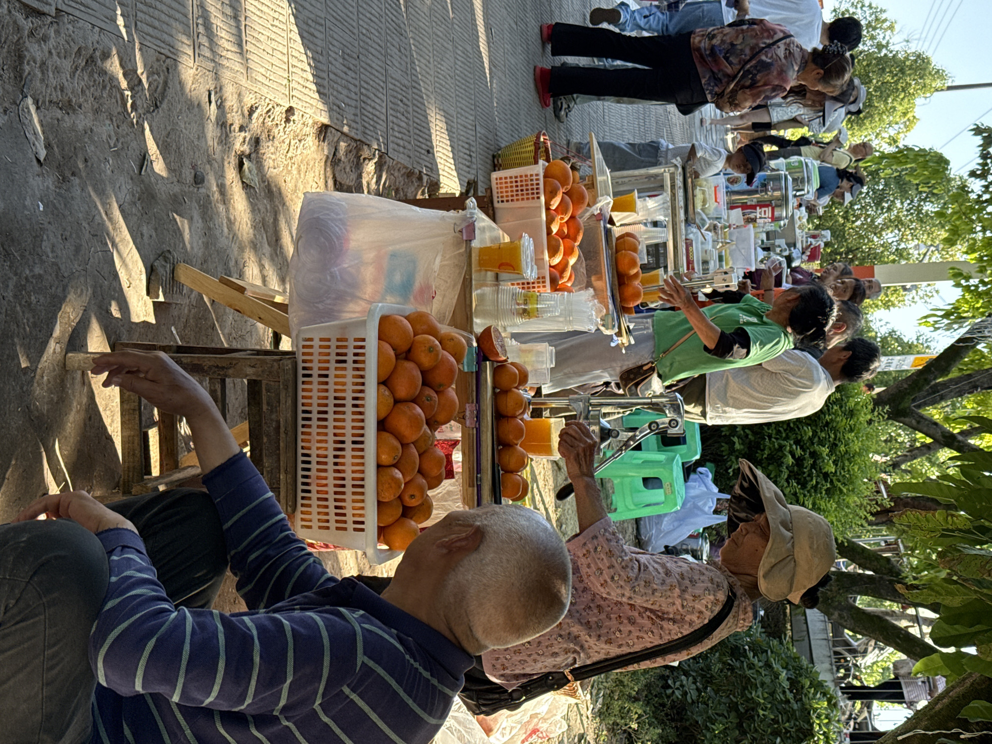The image shows a street scene in Beibei, China, where an orange juice vendor is serving customers. The vendor has set up a stall on the sidewalk, with crates of oranges, cups, and juice-making equipment. Several customers are lined up at the stall, waiting to purchase juice. Some are already enjoying their drinks. The setting is informal and lively, with people interacting with the vendor and each other. Trees and other foliage are visible in the background, suggesting the stall is located in a park or another area with greenery.