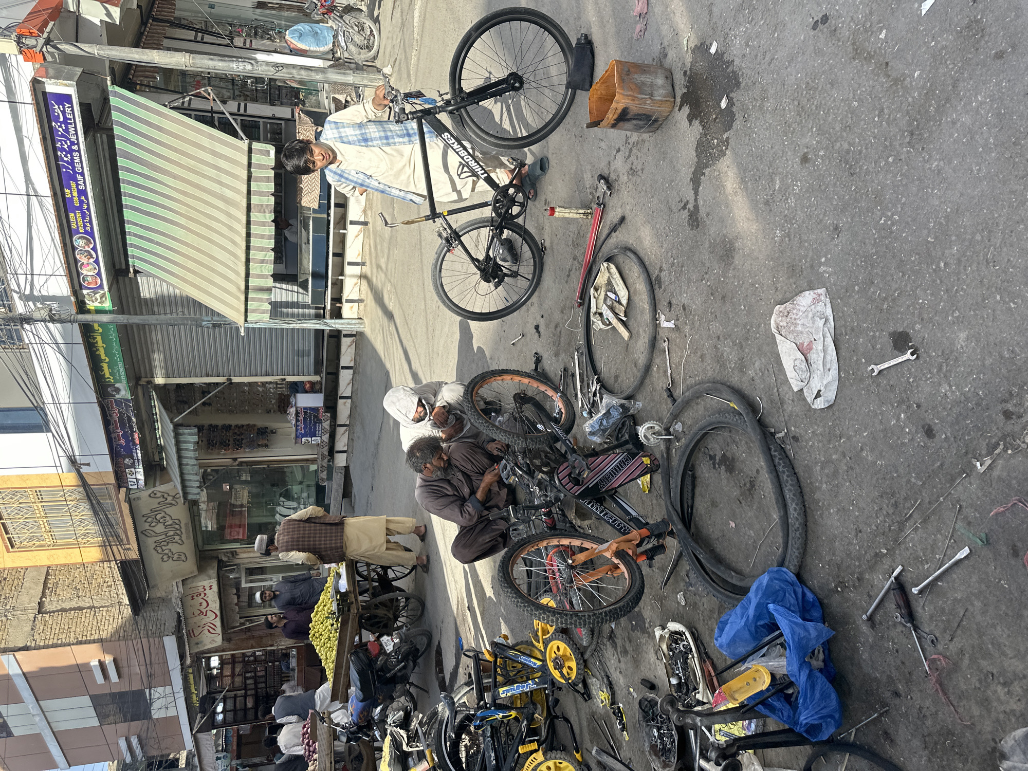 The image depicts a street scene in Quetta City Tehsil, Pakistan, likely a marketplace. In the foreground, two men are engaged in repairing bicycles. One man, wearing a brown tunic and cap, is hunched over a disassembled bicycle, working on its components. Another man, in a white hooded garment, leans in to assist him. Scattered around them are bicycle parts, tools, and a blue plastic bag. A partially assembled black bicycle stands upright to the right, with a young man in a checkered vest and shalwar kameez standing beside it. In the background, a market stall displays various items, including fruit, and other shops with awnings line the street. Motorbikes are parked along the side. The ground is paved with asphalt, showing signs of wear and some debris.