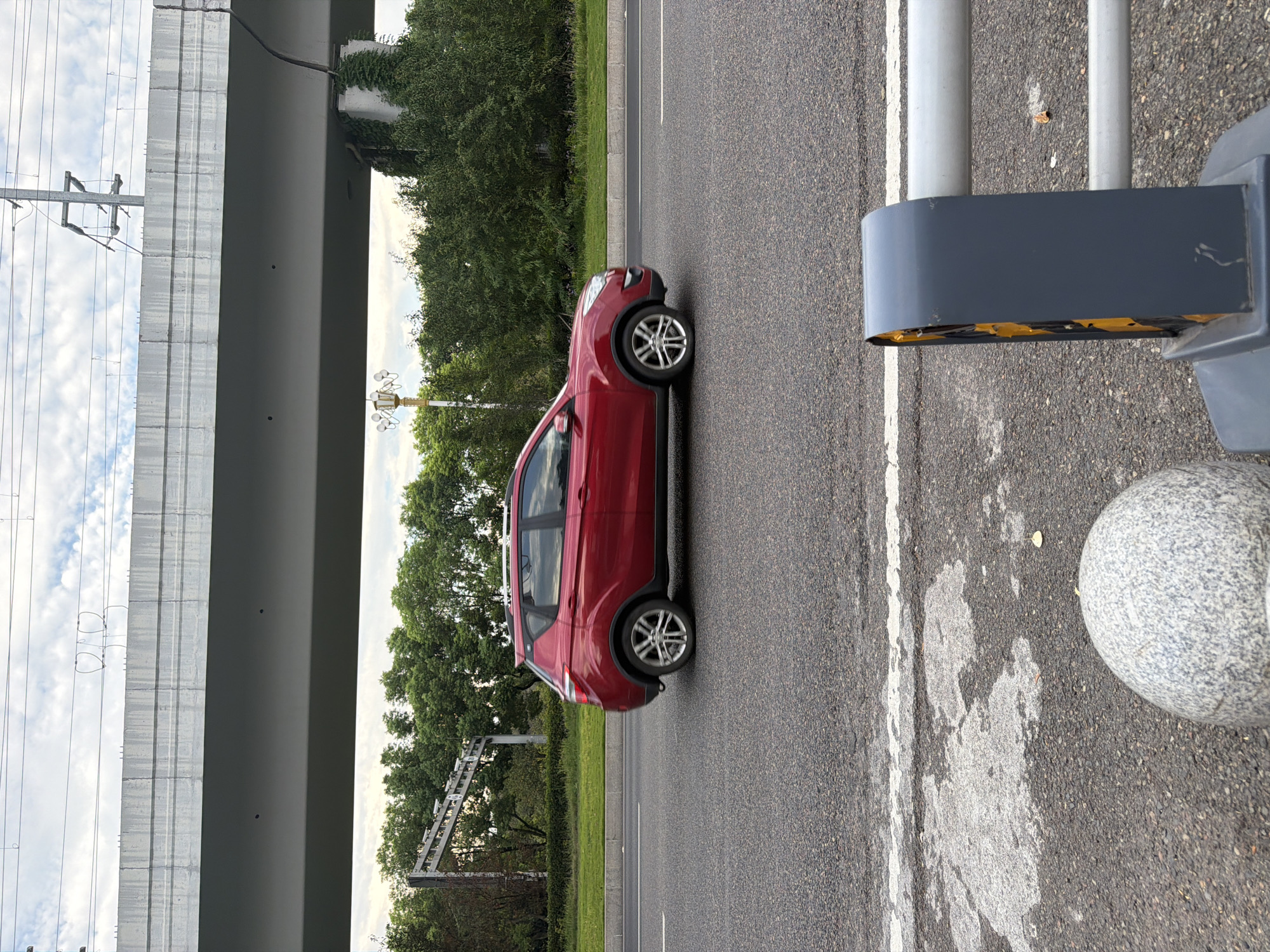 A red SUV is driving on a paved road in Jiangjin, China. The car is positioned in the left lane, moving from left to right across the frame. Above the road, a concrete overpass runs horizontally, supported by pillars. Electrical wires are suspended from poles above the overpass. On the right side of the frame, a grey barrier with yellow reflective tape is visible, along with a spherical stone bollard. The background consists of green trees and a cloudy sky. The road has white lane markings.