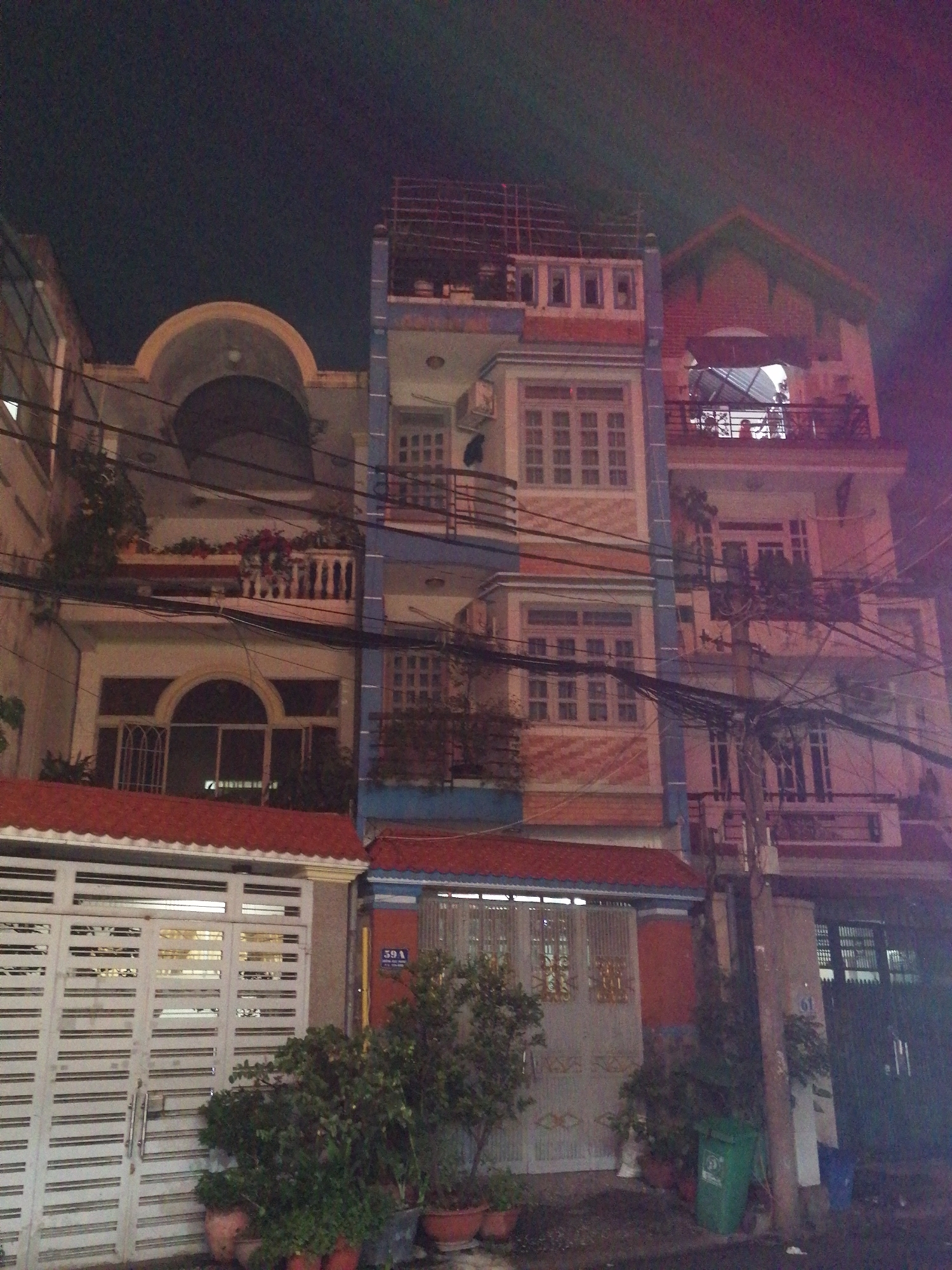 The image shows a row of multi-story buildings at night in Ho Chi Minh City, Vietnam. The buildings are narrow and tall, typical of the city's architecture. The facade of the buildings is a mix of pastel colors, light pink, and blue. Several windows and balconies are visible, some with plants and decorations. The street in front of the buildings is paved and there are trees and potted plants. A white metal gate is visible on the left side of the image. The sky is dark, indicating it is night time. Electrical wires are visible crisscrossing in front of the buildings. There is a sign on one of the buildings that reads "59A".