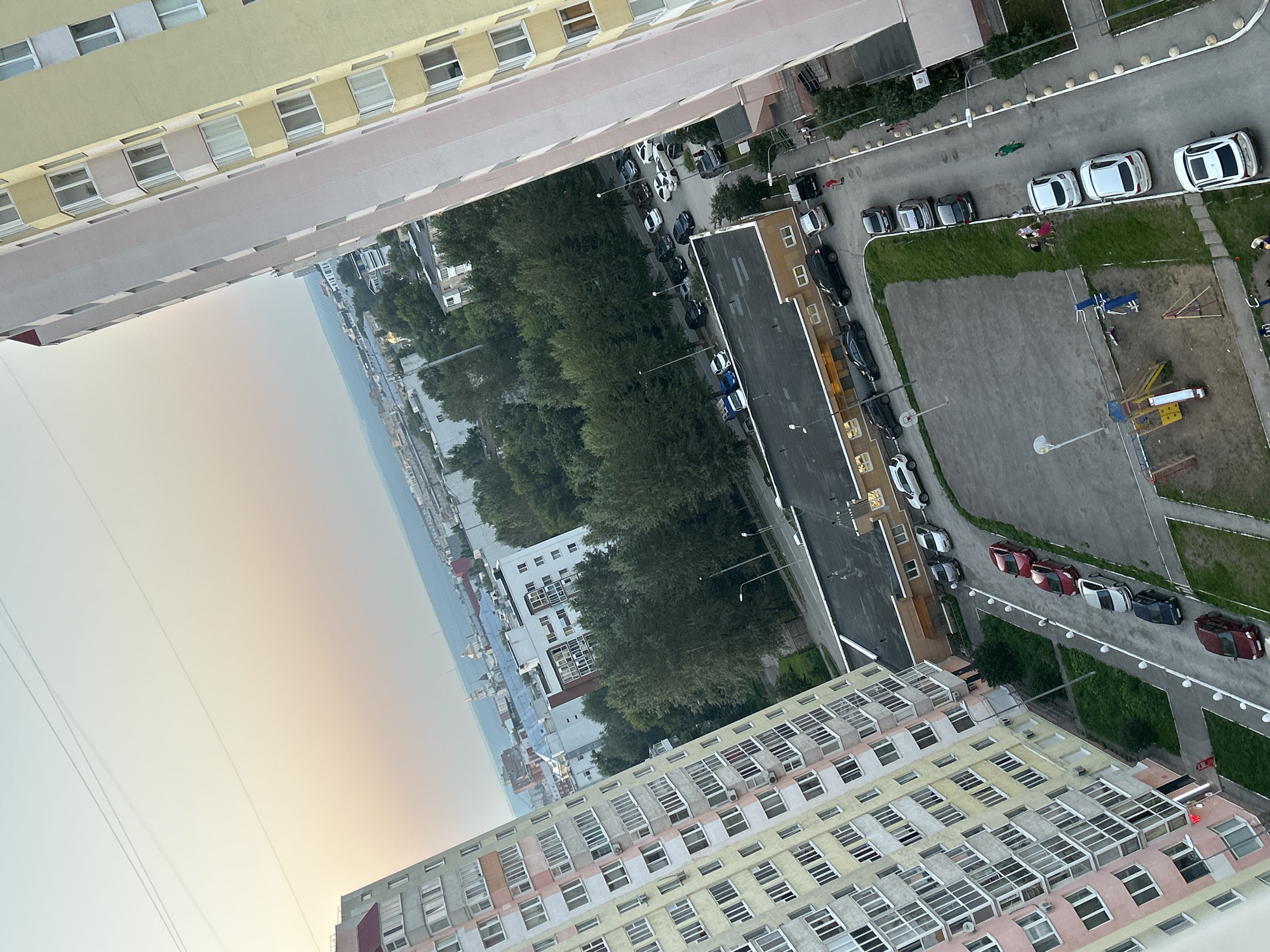 An aerial view captures a residential area in Perm, Russia, under a soft, hazy sky, possibly at dawn or dusk. Two high-rise apartment buildings dominate the foreground. The building on the left is lighter in color with pink and yellow accents and numerous balconies. The building on the right is taller and a pale yellow with white trim. Between these buildings, a low-rise building with a flat, dark roof is visible, adjacent to a parking area filled with various cars, including sedans and SUVs. Several trees line the streets and a small playground with slides and swings is present. People are sparsely distributed throughout the scene; a few individuals are walking on a sidewalk or near the playground. In the distance, other buildings and structures are visible, suggesting a larger urban environment, with a body of water and more distant landmasses extending to the horizon.