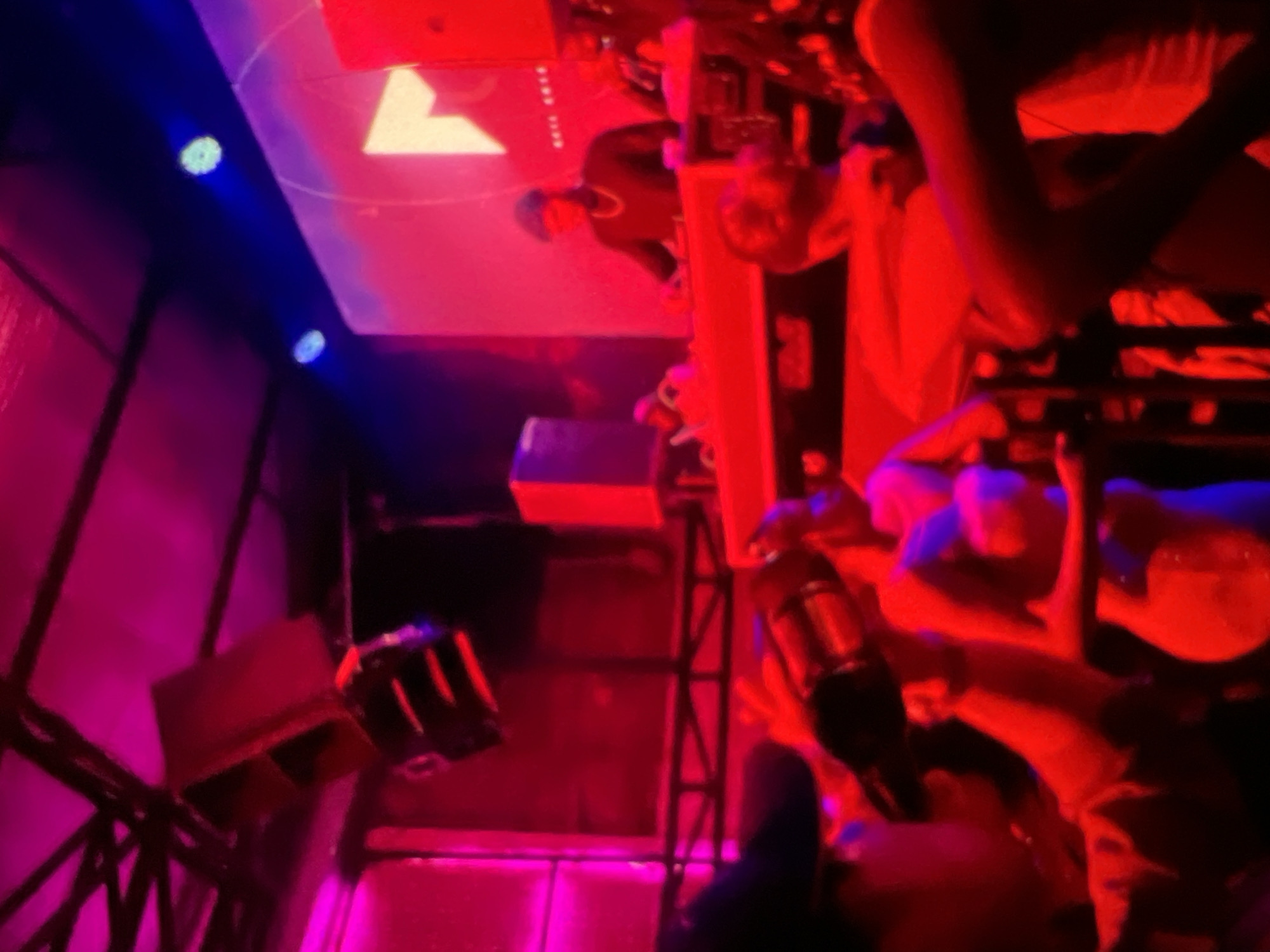 The image captures a dimly lit indoor venue, likely a club or performance space, saturated with vibrant red and pink lighting. In the background, a large screen displays a yellow geometric shape, possibly a logo, with a blue light source illuminating it from above. Suspended from the ceiling are black speakers.
In the mid-ground, a DJ, wearing a dark cap and a necklace, stands behind a console, appearing to be operating equipment. A few other individuals are visible in the vicinity of the DJ.
The foreground and mid-ground are populated with several people, most of whom are either blurred or partially obscured. One person in the foreground is holding a metallic object, possibly a bottle or a microphone, which is illuminated by the red lighting. Other individuals are engaged in various activities, suggesting a social gathering or a lively event. The overall impression is one of a dynamic, possibly musical, event taking place in a nightlife setting in Lagos, Nigeria.