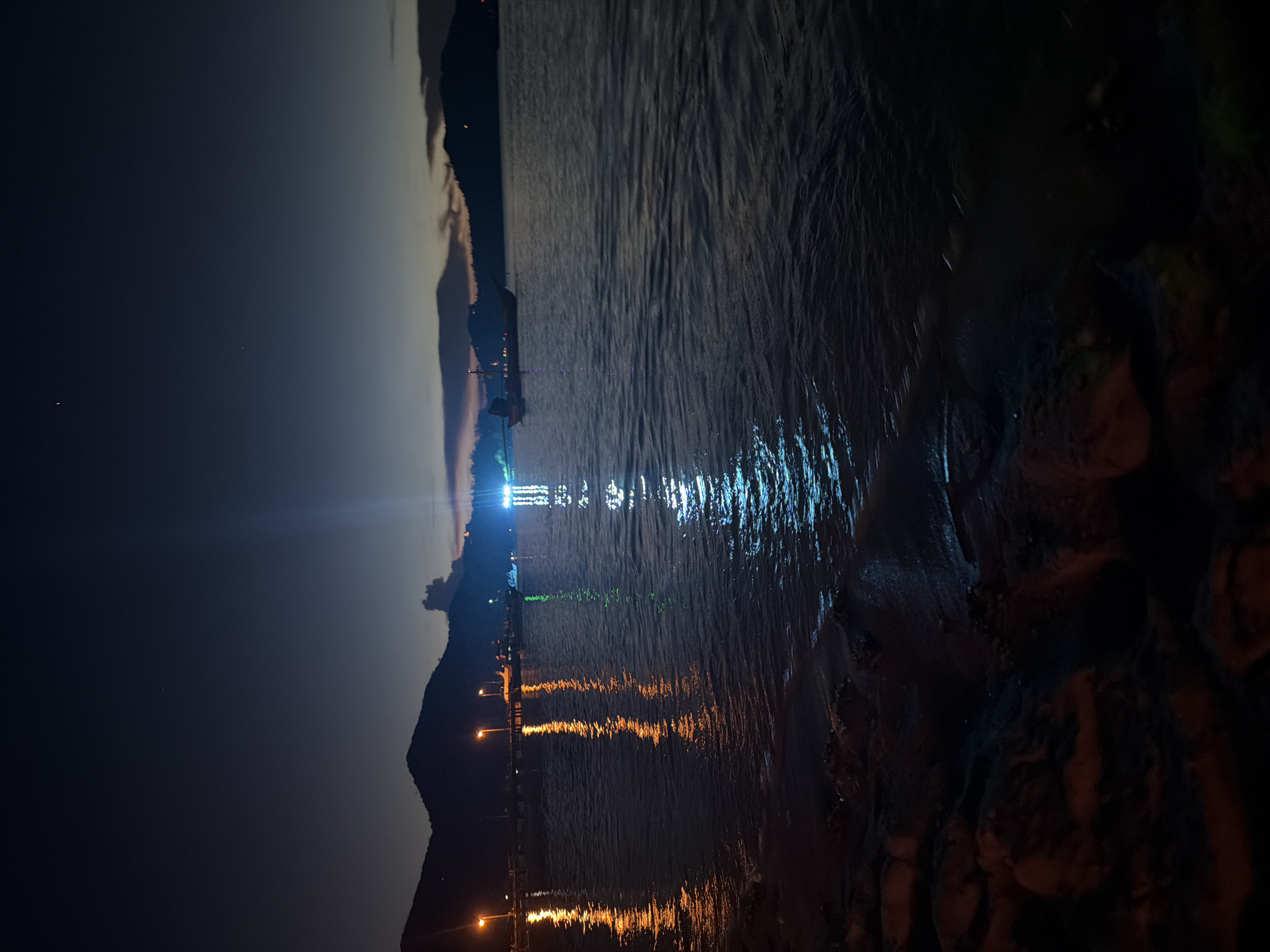 The image depicts a nighttime scene of a body of water, likely a bay or sea, with a mountainous coastline in the background. Several illuminated structures, possibly streetlights, are visible along the shoreline, casting reflections on the water's surface in shades of orange and yellow. In the middle ground, a boat with lights is anchored, and further out, other vessels and illuminated structures are faintly visible. The water itself has a gentle ripple, and the sky is a deep twilight blue fading to black. The foreground shows a rocky, dark shoreline. The scene is located in Koh Phangan, Thailand, specifically in Tambon Koh Phangan.