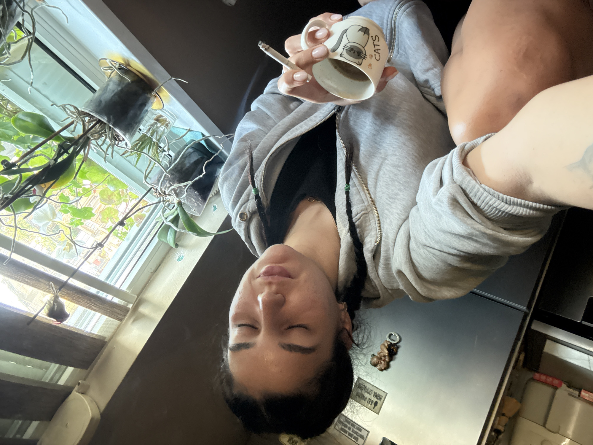 A woman with closed eyes holds a "CATS" mug and a lit cigarette indoors. She wears a gray hooded sweatshirt and has dark hair. A window with plants is to her left. A refrigerator is to her right, with magnets on the door. The location is unknown, and there is no indication of Veliko Tarnovo, Bulgaria.