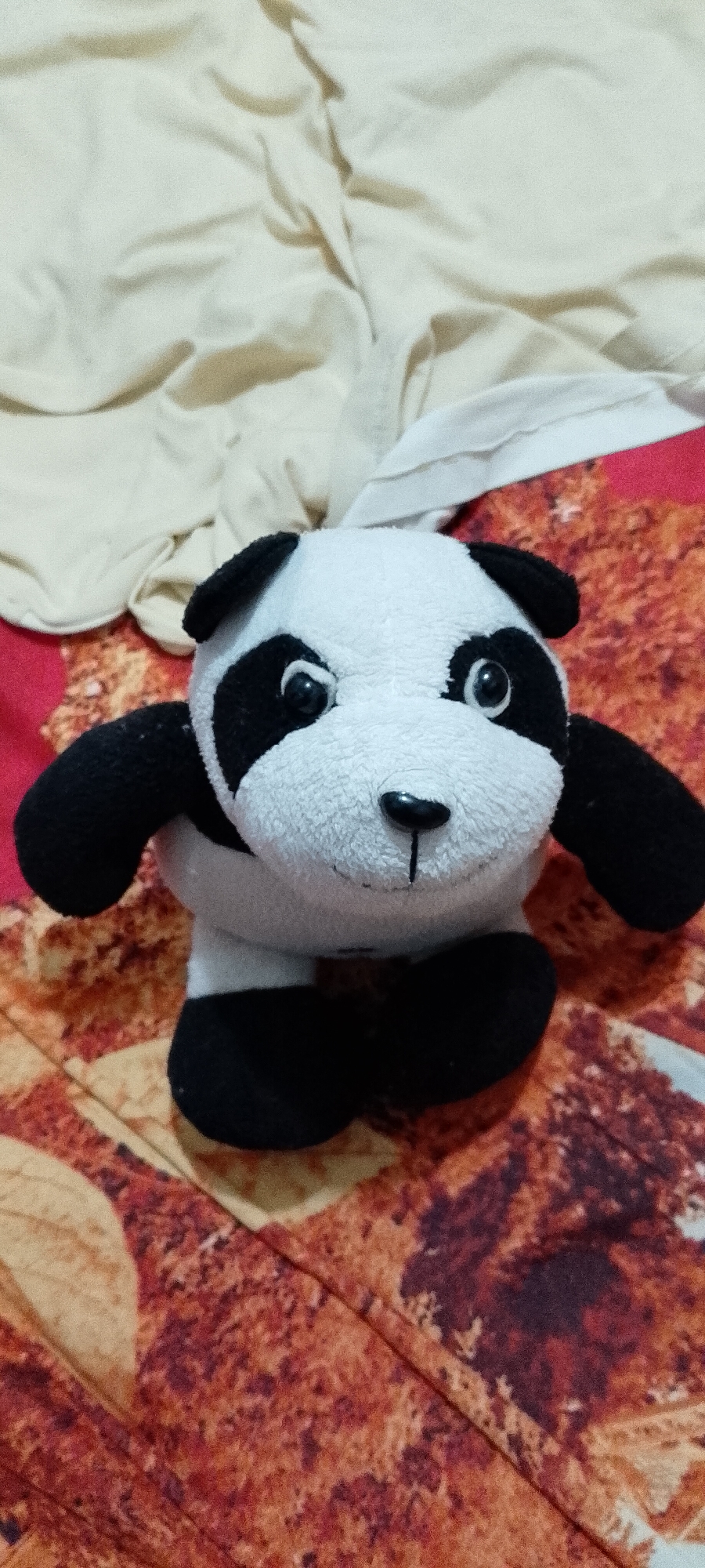 A stuffed panda toy is positioned upright on a patterned surface, which appears to be a bedspread with reddish-brown and tan motifs. The panda is black and white, with black patches around its eyes and ears. It has a black nose and mouth, and its eyes are black. The background consists of a light-colored, rumpled fabric, possibly a blanket or sheet, draped behind the toy. The lighting suggests an indoor setting. There are no people or visible human interaction in the image. There is no discernible information within the image that would indicate its location in Singkawang, Indonesia.
