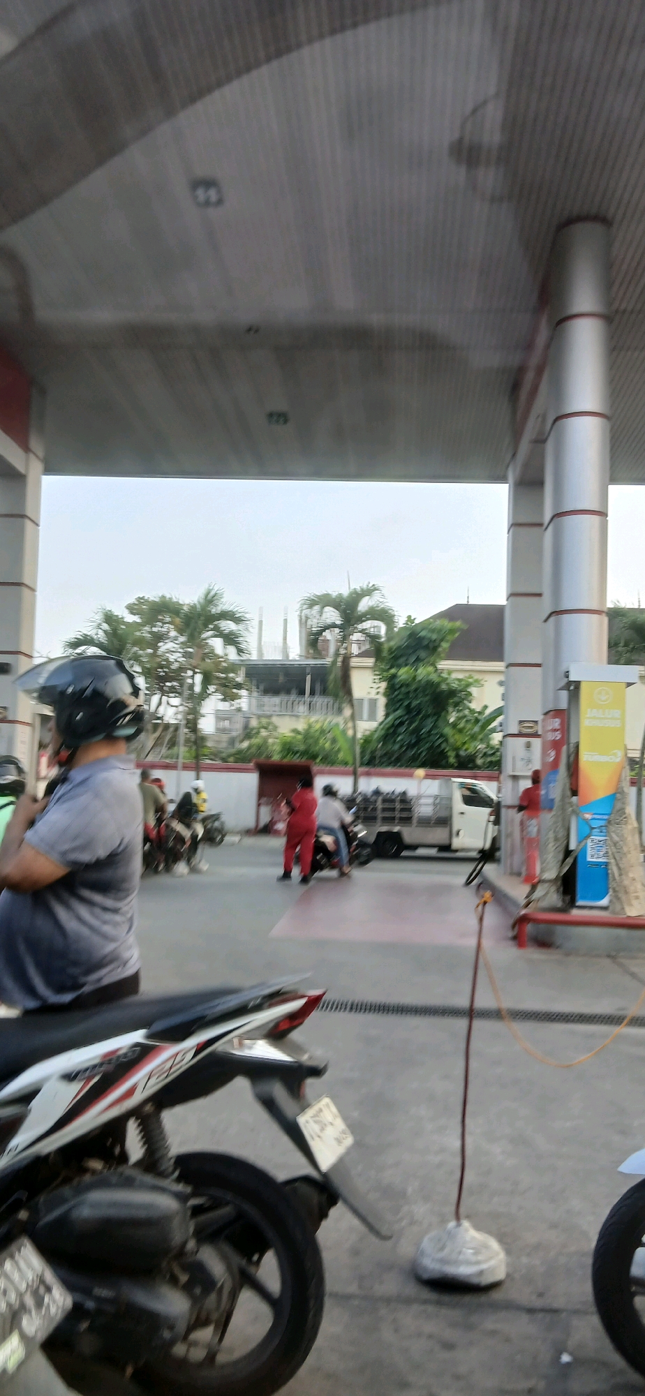 The image captures a scene at a gas station, likely in Samarinda, Indonesia, given the visual cues. In the foreground, a person wearing a gray t-shirt and a black helmet is partially visible, with their back to the camera, looking towards the gas station. A portion of a white and black motorcycle is also in the immediate foreground, with its rear wheel and fender clearly depicted.
The gas station itself features red and white striped pillars supporting a large, sloped roof structure. Several motorcycles are present in the middle ground, some with riders. One person is wearing a bright red uniform and appears to be attending to something at the gas pumps. Another person in a light-colored shirt is on a motorcycle. A white utility truck with a caged bed is parked near the pumps.
In the background, palm trees and a building under construction are visible under a hazy sky. A sign with the word "JALUR KHUSUS" is displayed on one of the gas station pillars, indicating a "special lane" or "dedicated lane," a common feature at Indonesian gas stations. The ground is paved, and a dark drainage grate runs horizontally across the mid-ground. The overall lighting suggests it is daytime, possibly late afternoon.