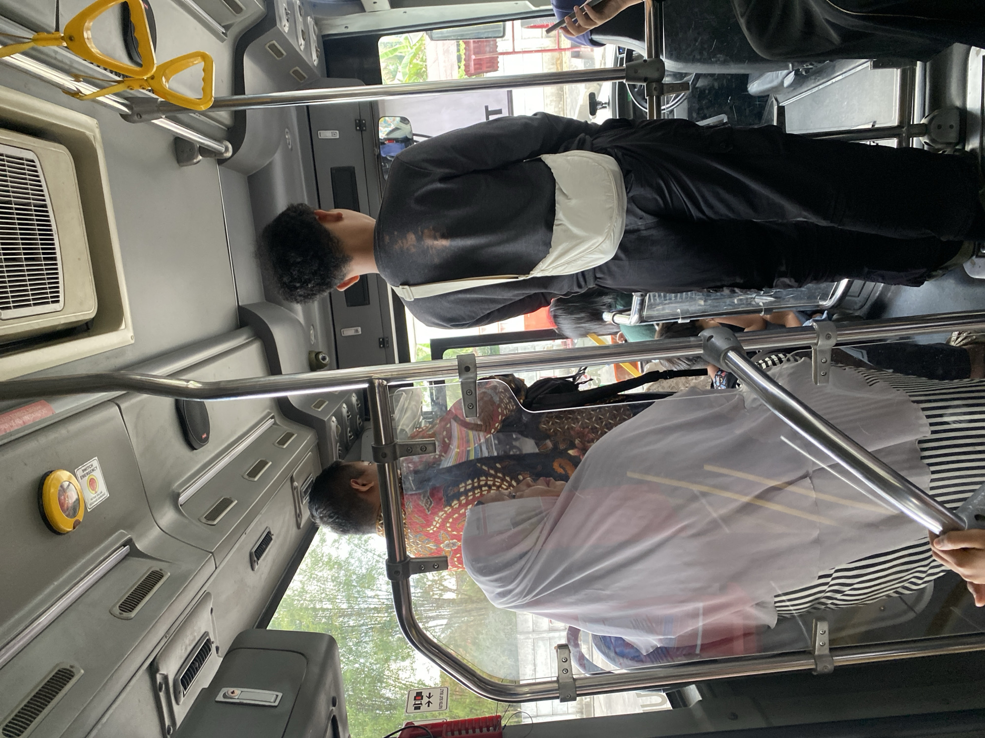 The image shows the interior of a bus, possibly a public transport bus, in Banjarbaru, Indonesia. The perspective is from inside the bus, looking towards the front. In the foreground, to the left, a person wearing a white hijab and a striped skirt is partially visible, holding onto a metal railing. Behind them, a person wearing a patterned red and black shirt is standing. In the center of the frame, a young person with curly hair is standing, wearing a dark sweatshirt and black pants, with a white crossbody bag slung over their shoulder. The bus interior features metal poles, yellow hand straps hanging from the ceiling, and air vents. The environment outside the bus, visible through the windows, appears to be green foliage, possibly trees, and a concrete wall. The lighting suggests it is daytime.