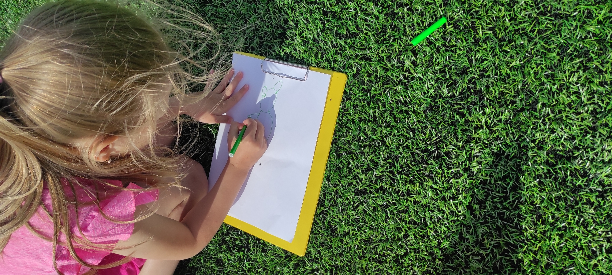 A young girl with blonde hair, wearing a pink shirt, kneels on artificial grass. She is drawing on a white sheet of paper secured to a yellow clipboard. A green marker is in her right hand, actively drawing. Another green marker lies on the grass to the lower right of the image. The scene is lit by sunlight, creating shadows on the grass. The location of Noviy, Russia, is not discernible from the image.