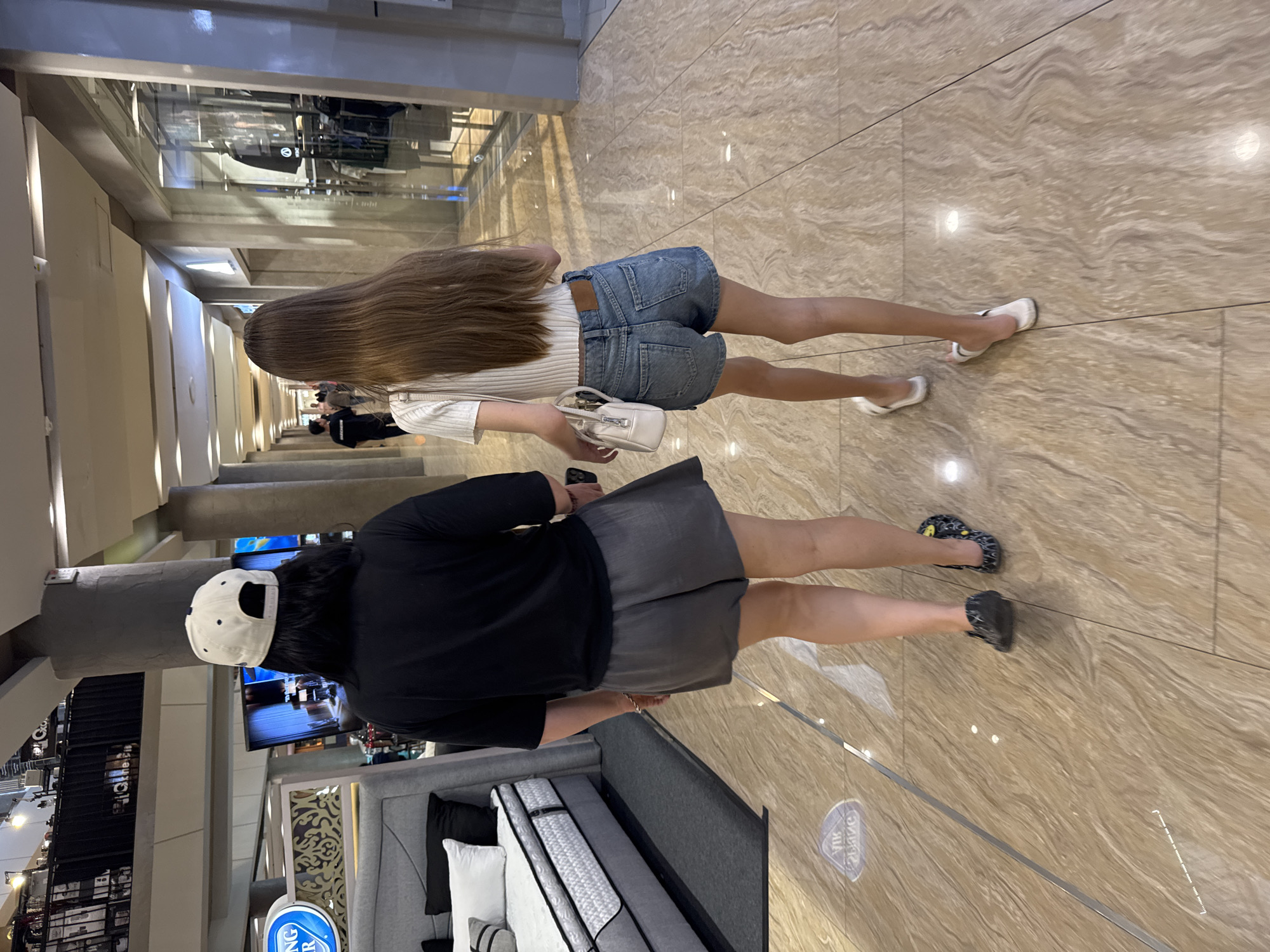 Two women are walking through a shopping mall. One is wearing a white top, denim shorts, and white sandals, carrying a small white purse. The other is wearing a black top, gray skirt, and black shoes. A white baseball cap is on her head. They are walking on a marble-tiled floor. The location may be in the city of Denpasar, Indonesia. Beds and other merchandise can be seen in the background, indicating they are near a store.