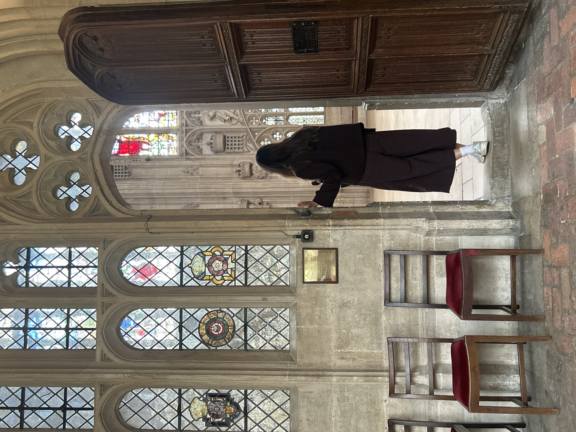 An interior shot shows a person standing in a doorway of a large stone building in Cambridge, United Kingdom. The person, viewed from behind, is wearing dark clothing and white sneakers, and has long, dark hair. They are looking into a room with stained glass windows and intricate stone architecture. To the left of the doorway, two wooden chairs with red velvet seats are visible. The overall impression is of a historical or religious setting, possibly within a college or cathedral. The lighting is subdued, with natural light filtering through the stained glass.