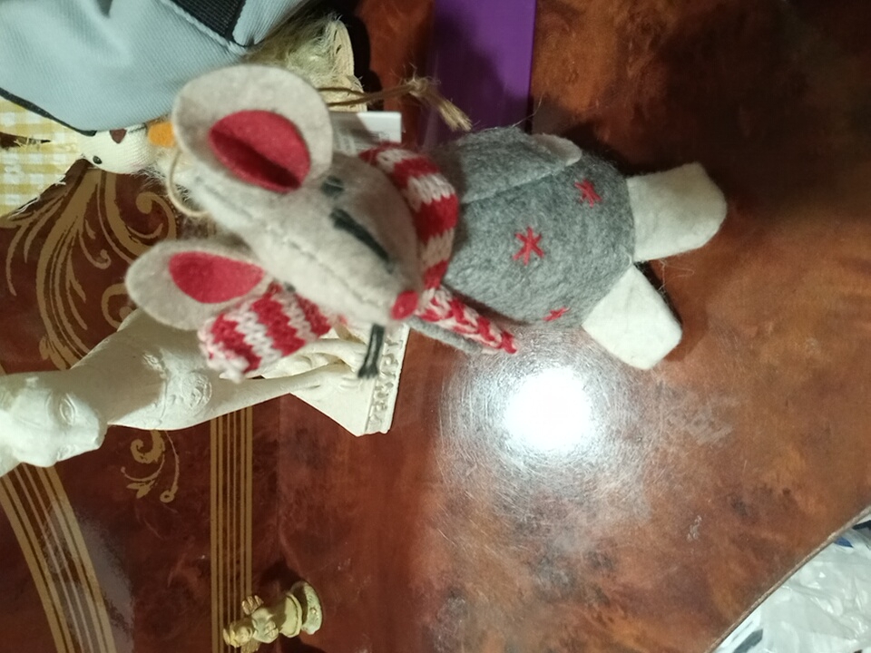 In Angren, Uzbekistan, a close-up, eye-level, indoor photograph captures a wooden table surface. On the table, a small, gray, felt mouse ornament with red accents is positioned in the foreground, slightly off-center to the right. The mouse is wearing a red and white striped scarf and has red stitched crosses on its chest. White felt feet extend from its body. Behind the mouse, a white, stylized statue of a cat with hieroglyphics at its base is visible. To the far left, a miniature angel figurine stands. In the background, a portion of a blue bag and a blurry white and yellow patterned object are discernible. The lighting suggests an indoor environment, possibly a home setting.
