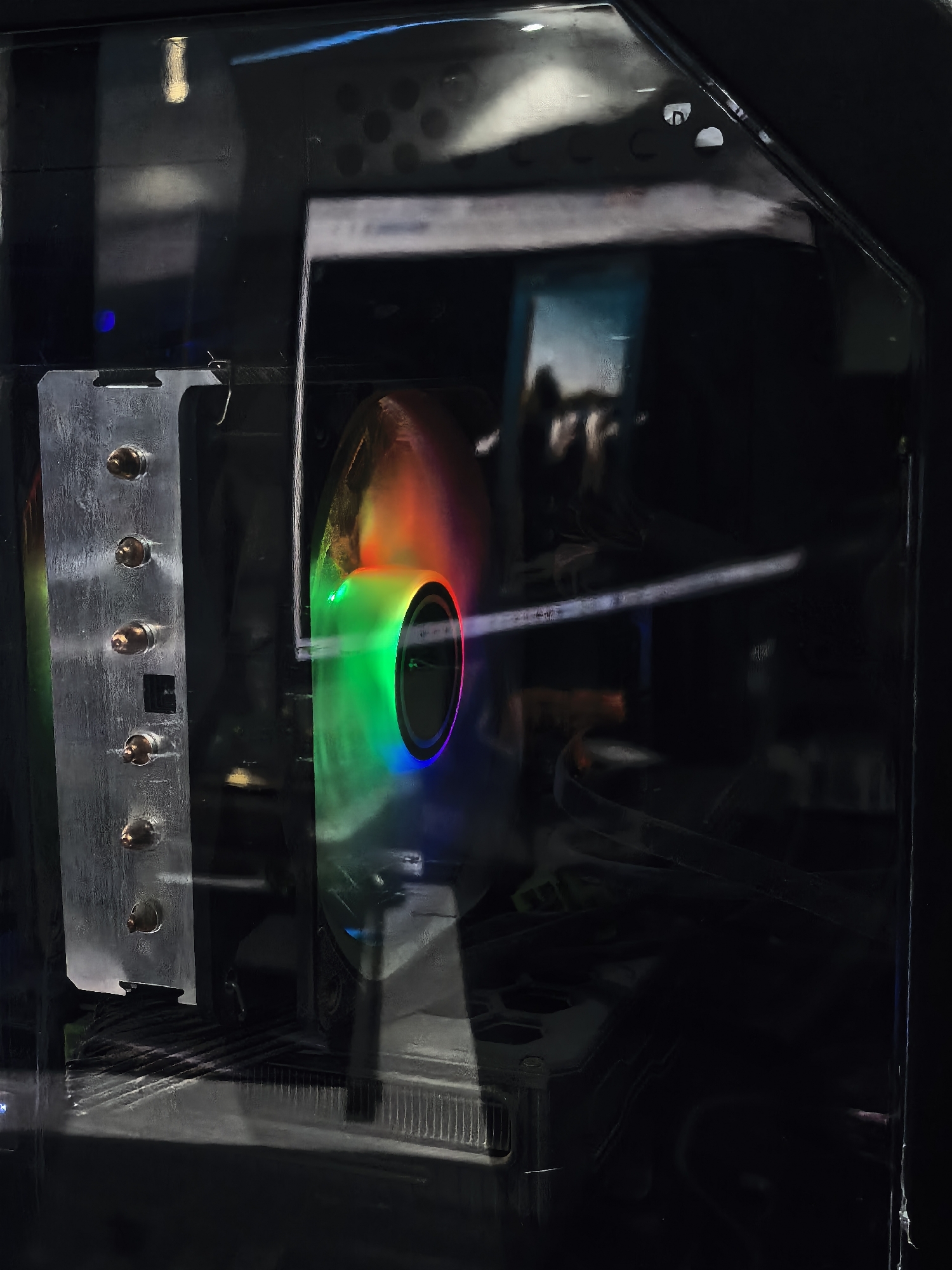 The image shows a close-up of a computer's internal components through a transparent casing. A CPU cooler with colorful LED lights is prominently featured, showing a gradient of red, green, and blue. To the left of the cooler, a metal heatsink with visible copper heat pipes is attached. The background is dark and somewhat out of focus, with hints of other computer hardware and cables. The overall impression is one of a modern, customized computer build. The image does not contain any information that would allow for its localization in Fergana City, Uzbekistan.