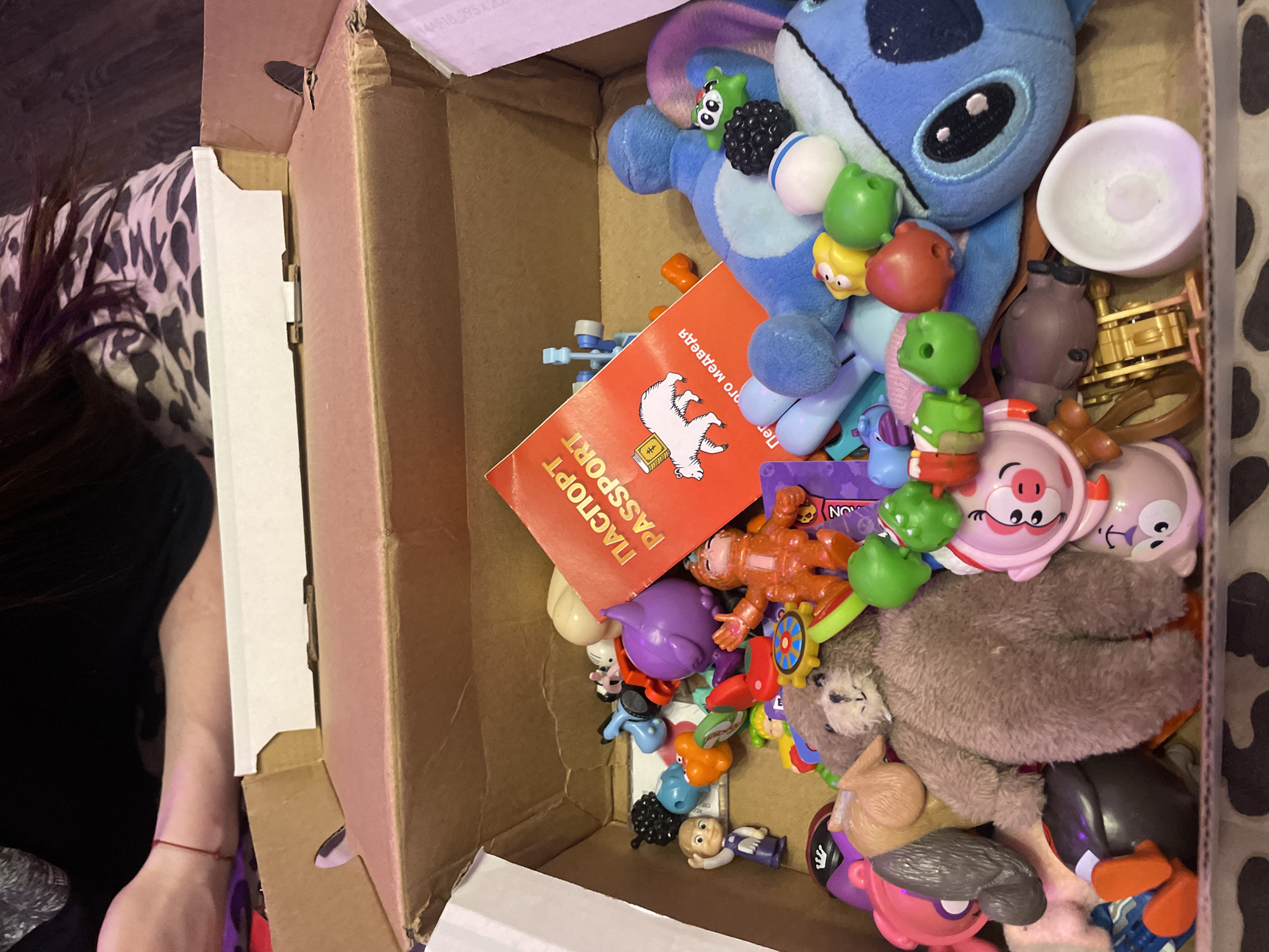 A box of toys sits on a patterned surface. The box is open, revealing a jumble of various toys, including plush figures, small plastic toys, and a red passport-like booklet. A person's arm, with a red string bracelet, is visible at the top left of the image. The background appears to be indoors, with hints of furniture and a patterned fabric. The scene is likely a domestic setting. The location of Kokkorevo, Russia is not determinable from the image.
