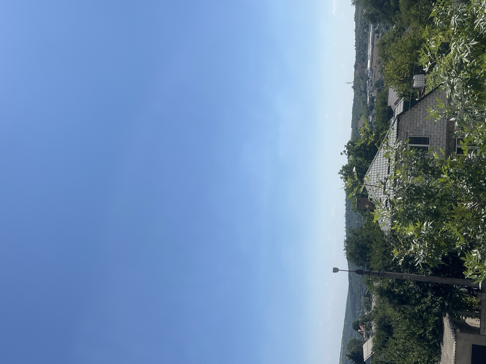 The image shows a clear blue sky over a rural landscape, likely on the outskirts of Rybnitsa, Moldova. In the foreground, a street lamp stands tall, partially obscured by lush green foliage. Several houses with tiled roofs are visible amidst dense trees, with one house constructed of white brick being prominent. In the middle ground, rolling hills and scattered buildings can be seen, suggesting a semi-urban or agricultural area. The overall impression is one of a peaceful, sunny day in a village setting.