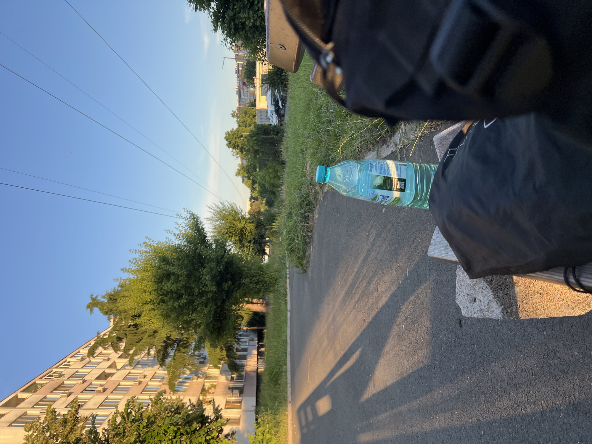 The image depicts an outdoor scene with a residential building on the left, tall green trees, and a clear blue sky. In the foreground, a paved pathway is visible, casting long shadows from the setting sun. A black backpack is placed on a wooden bench on the right side of the frame, with a partially visible bottle of mineral water next to it. The overall ambiance suggests a tranquil moment in an urban environment, possibly a park or a public space. The building, characterized by its uniform balconies and windows, is typical of socialist-era architecture found in many Eastern European cities, including Burgas, Bulgaria. The presence of power lines overhead also points to an urban setting. The vegetation and the quality of light suggest it is late afternoon or early evening during a warm season.