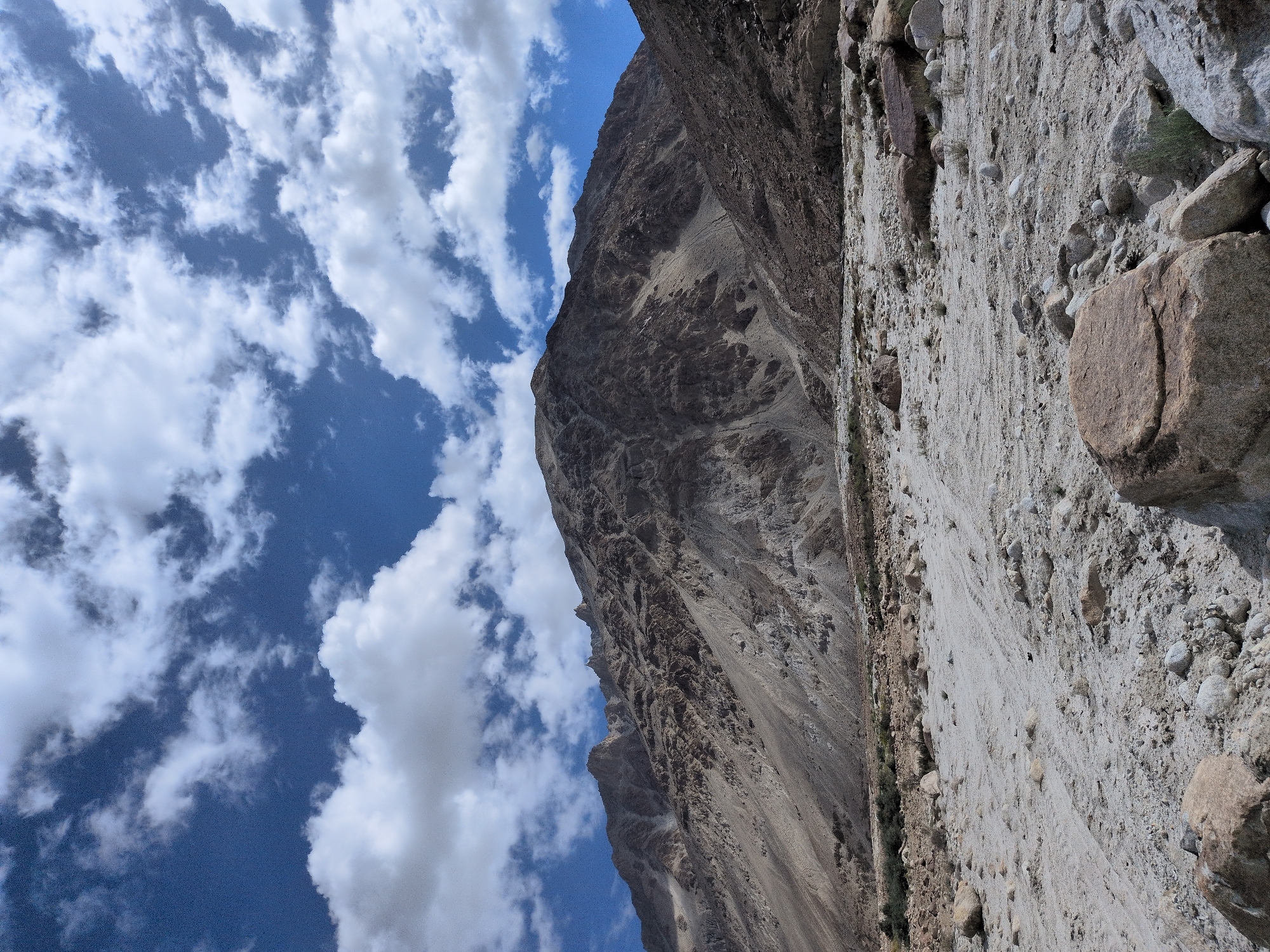 The image depicts a vast, rocky, and sandy terrain with a mountain range dominating the background. The sky above is a vibrant blue, dotted with scattered white clouds. In the foreground, numerous stones and boulders of varying sizes are strewn across the dry, light-colored ground, which shows faint tire tracks. The mountain slopes are rugged and arid, with visible geological layers and shadows suggesting uneven terrain. The scene appears to be a natural landscape, likely in a mountainous region. There are no people or man-made structures visible in the image. The location is identified as Sasoma, India.