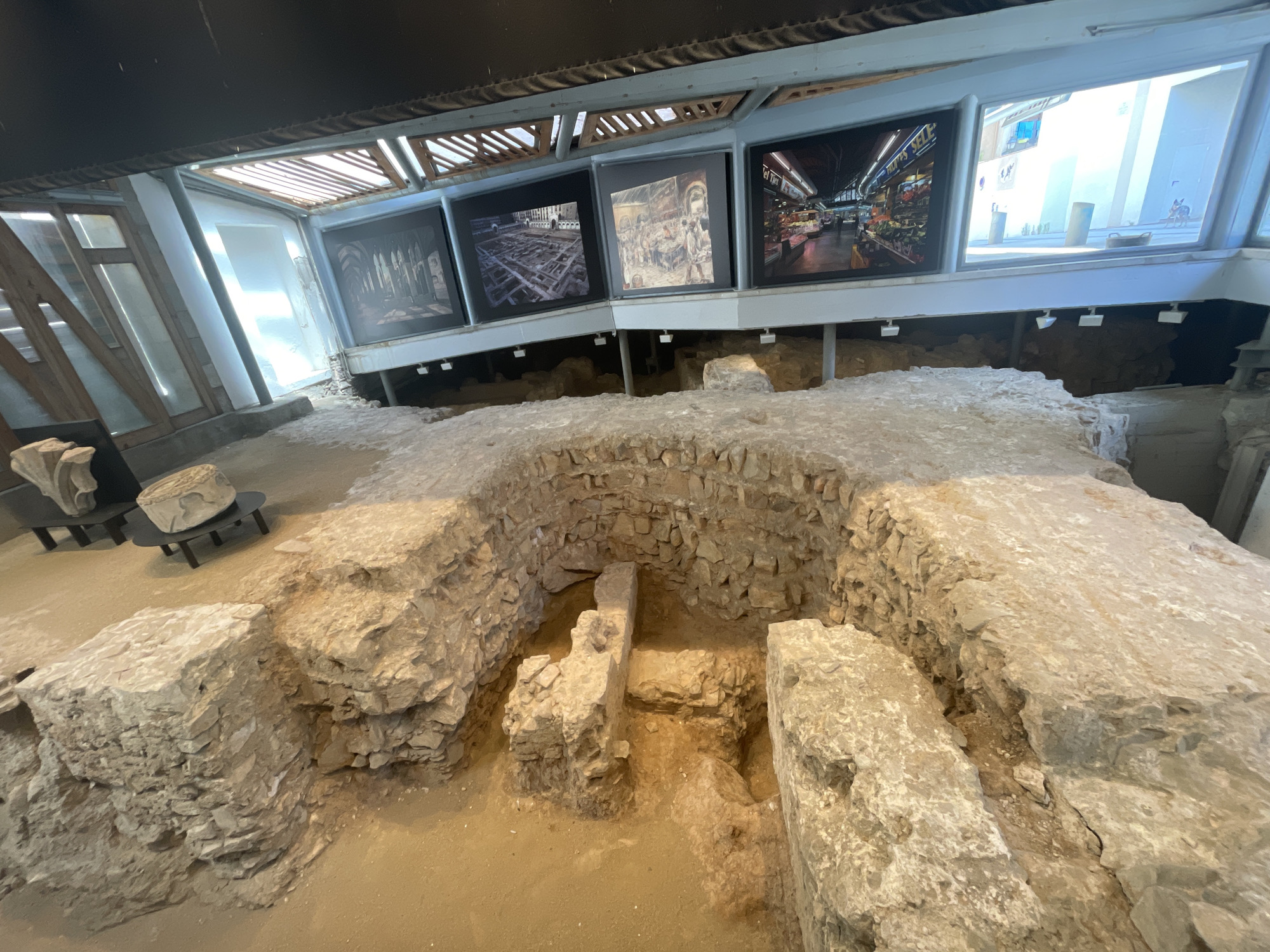 The media file presents an indoor archaeological exhibit, likely situated in Barcelona, Spain. The central and foreground areas are dominated by excavated ancient stone ruins, revealing foundations and walls constructed from rough-hewn blocks. A prominent, deeper pit with stone walls is visible in the midground, suggesting further architectural complexity. The ground surrounding these ruins is composed of sand or earth.
Above the archaeological site, a modern structure provides an enclosure and display area. This structure features large windows and an array of horizontal display screens mounted on an elevated platform. Four distinct images are shown on these screens: from left to right, an interior view of an ancient arched hall, an aerial perspective of a historical site excavation, an artistic depiction of a lively market scene with figures, and a modern photograph of a vibrant supermarket aisle filled with produce. Faint text, possibly "MERCAT DE...", is visible on one of the market-themed screens. Beyond the screens, large windows offer a view of a brightly lit area, possibly an outdoor street or courtyard, characterized by white buildings, a blue window, and a small, stylized donkey figure.
The exhibit is illuminated by multiple white spotlights positioned beneath the display screens, casting light onto the ruins below. To the far left, two black stands showcase additional stone artifacts or carved fragments. The scene conveys a sense of historical preservation and educational display, merging ancient discoveries with contemporary presentation methods. No people are visible in the image.