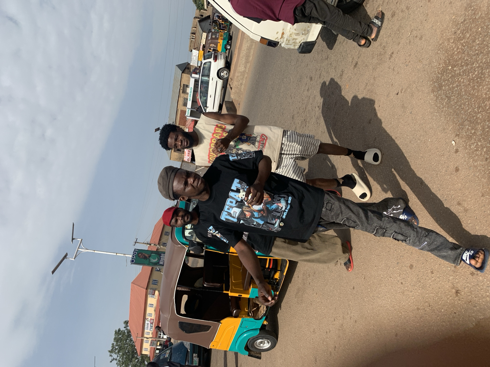 In Gura Top, Nigeria, on a paved road under a hazy sky, three young men are captured in a candid moment near a brown and yellow auto-rickshaw. The man in the foreground, wearing a dark t-shirt with a graphic print and distressed denim pants, holds a small object in his right hand and looks towards the camera. To his left, a man with a red beret and a similar t-shirt stands by the auto-rickshaw. Behind them, another man in a sleeveless t-shirt and striped shorts also looks ahead. The background shows buildings, a street light with solar panels, and other vehicles, including a white minivan. The scene is illuminated by natural daylight, casting shadows on the ground.