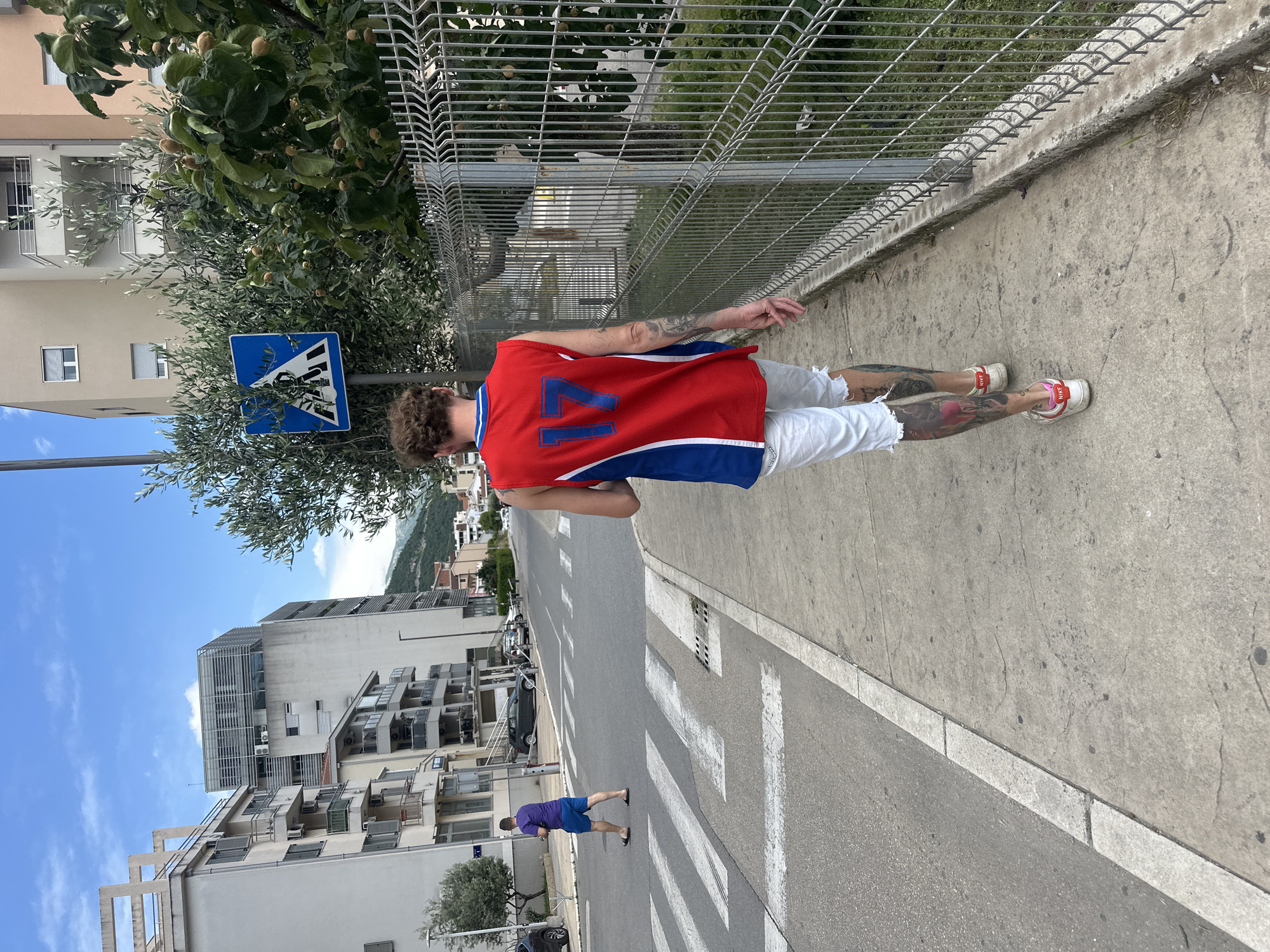 The photograph depicts a male standing on a sidewalk in Budva, Montenegro. The man is wearing a red, white, and blue basketball jersey with the number "77" on the back, white shorts, and sneakers. He has tattoos on his arms and legs. The sidewalk is adjacent to a road with a pedestrian crossing. In the background, there are buildings, a pedestrian crossing sign, trees, a metal fence, and the sky. Another pedestrian is seen crossing the street. The weather appears to be sunny.