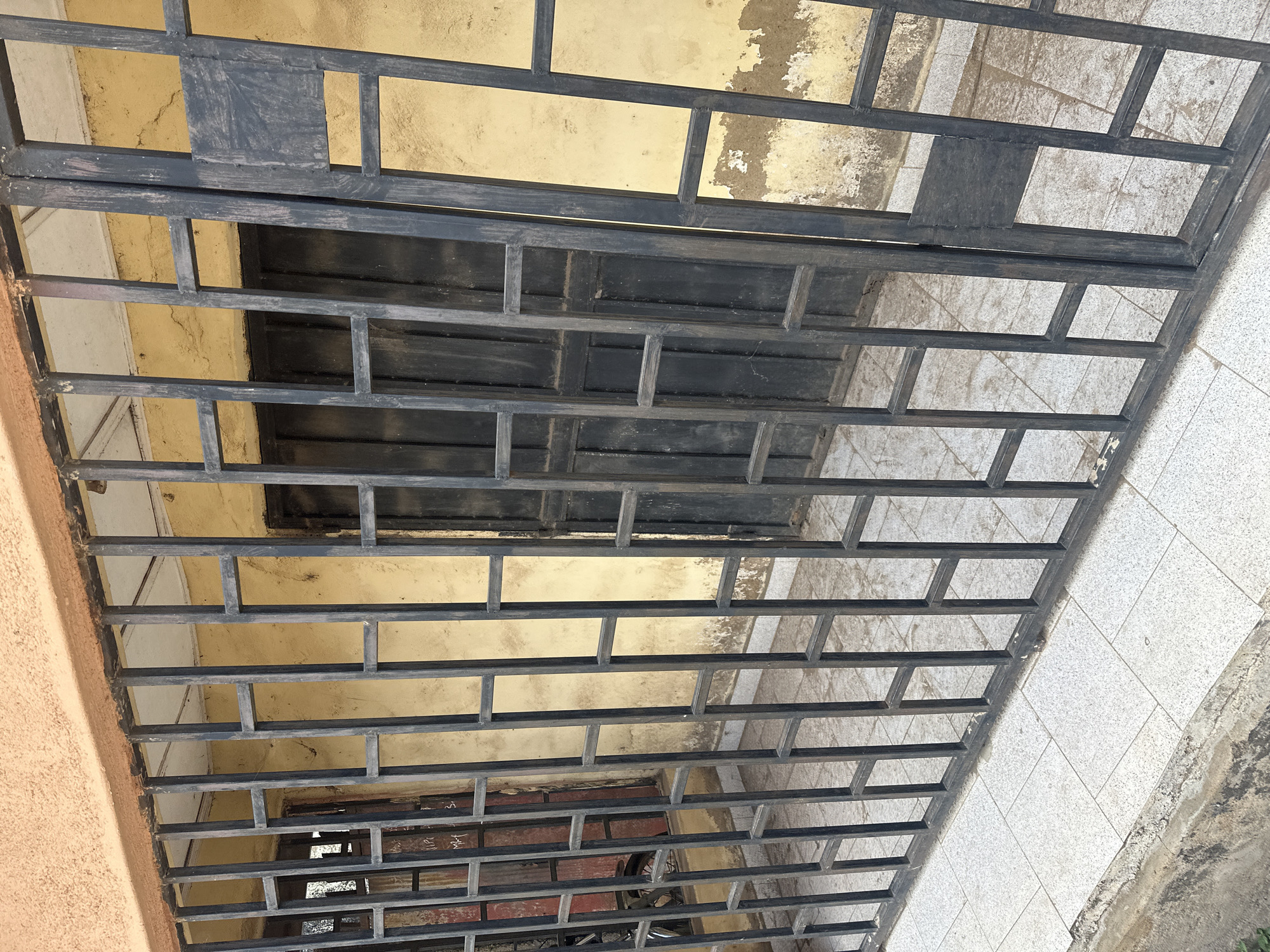 In an outdoor scene, a black metal grate is in the foreground, obscuring a building's entrance. The grate has a grid pattern with vertical bars and horizontal supports. Behind the grate, a yellow-painted wall shows signs of wear and peeling paint. Two large, dark metal doors are visible through the grate, likely serving as the main entrance. The ground in front of the building is paved with light-colored square tiles. The building has a textured orange overhang above the wall. The overall scene suggests a weathered urban environment.
