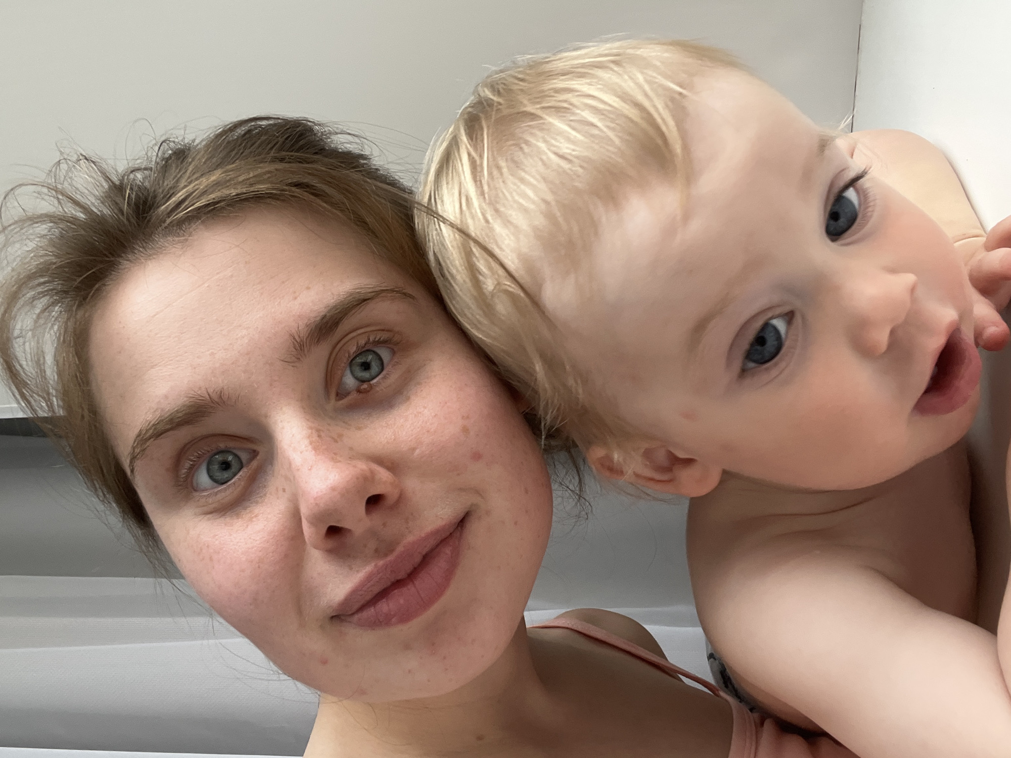 An indoor portrait, specifically a close-up selfie, depicts an adult female and an infant, taken in a domestic setting in the city of Chaikovsky, Russia. The adult, positioned in the upper portion of the frame, is a female with light skin, prominent freckles, and minor facial blemishes, possessing light blue eyes and light brown, somewhat disheveled hair. She wears a light pink/peach spaghetti strap garment and exhibits a faint, closed-mouth smile, with her head tilted. The infant, positioned in the lower portion of the frame, features light skin, light blonde hair, and light blue eyes, with its mouth slightly open. The adult's head is in close proximity to the infant's, suggesting an intimate interaction. The background consists of plain, light-colored surfaces, including a vertical light grey panel or curtain visible in the upper left quadrant.