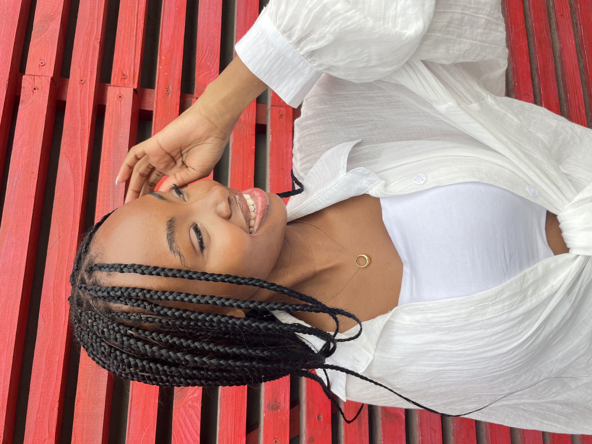 A young Black woman with braided hair is looking to her right and smiling, with her left hand touching her hair. She is wearing a white tube top and an unbuttoned white crinkled blouse. A thin gold chain with a circular pendant is around her neck. She is standing in front of a red slatted wooden wall. The location is not Kazan, Russia.