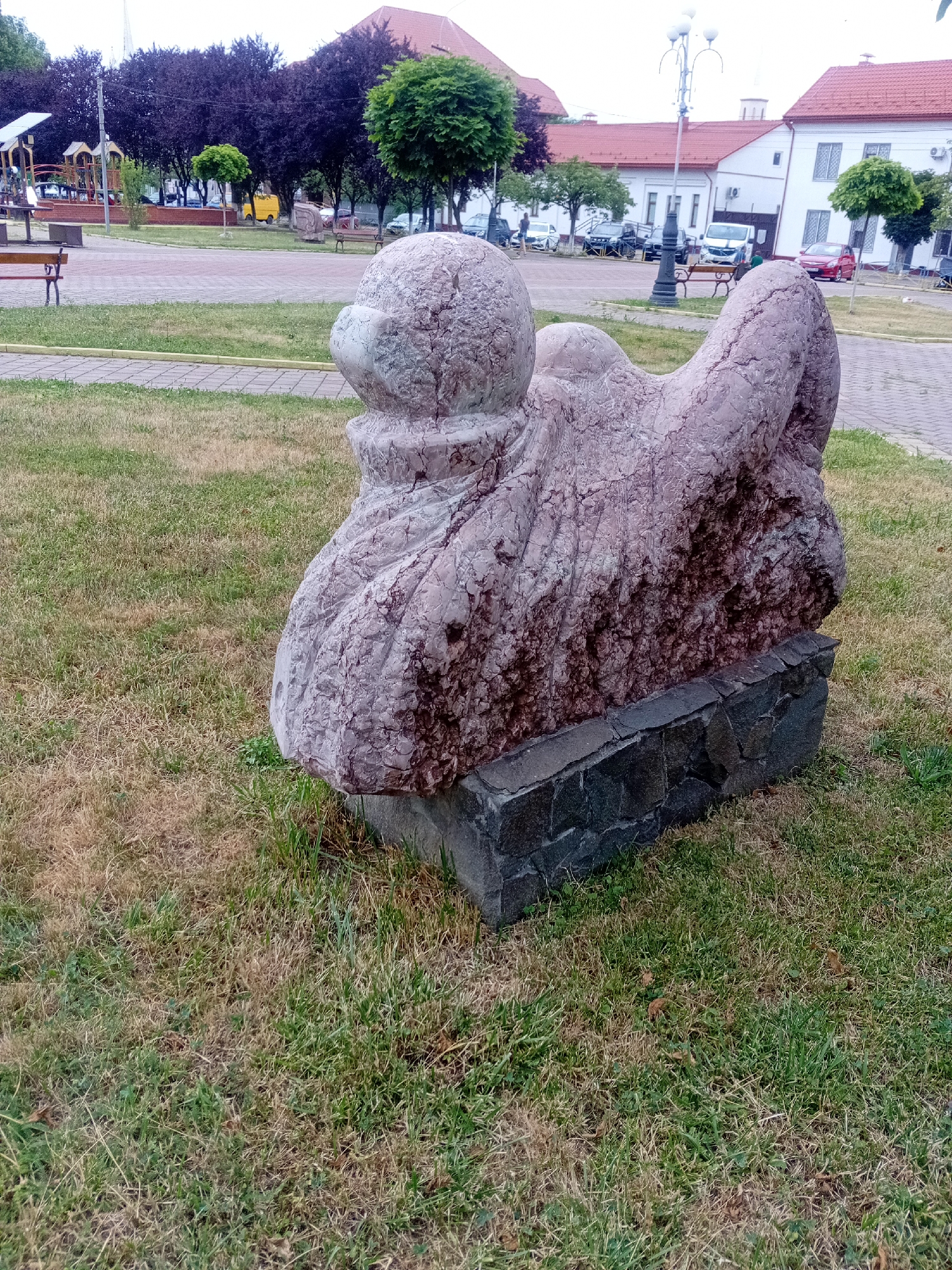 In a public park in Tiachiv, Ukraine, a large, abstract stone sculpture is the central subject. The sculpture, made of pinkish-purple and greyish rock with visible veins and rough textures, rests on a dark grey stone base. The sculpture appears to depict an organic, possibly animalistic form with rounded elements and a textured surface. The park setting includes a well-maintained lawn with areas of dry, brown grass interspersed with green. In the background, a paved walkway or plaza is visible, with benches and trees lining its edge. Further back, a playground structure with slides and climbing frames can be seen, along with parked cars and buildings with red-tiled roofs. The overall environment suggests a pleasant, urban green space.