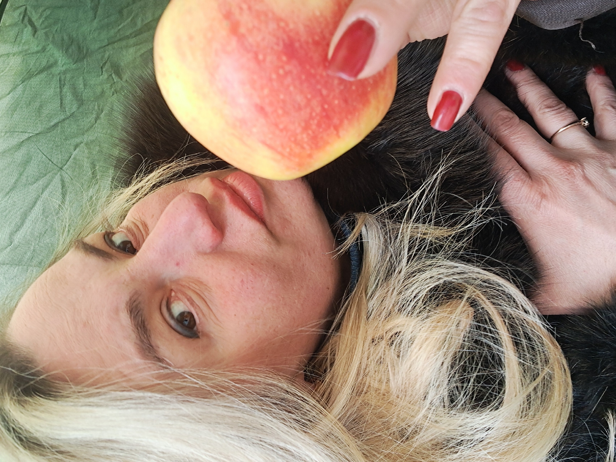 A woman with fair skin, blonde and dark brown hair, and red nail polish is holding a red apple near her face. She is wearing a dark-colored garment that appears to be fur-lined. The background is a green fabric. The image is taken at an unusual angle, with the woman appearing to be sideways. The location is not discernible from the image.