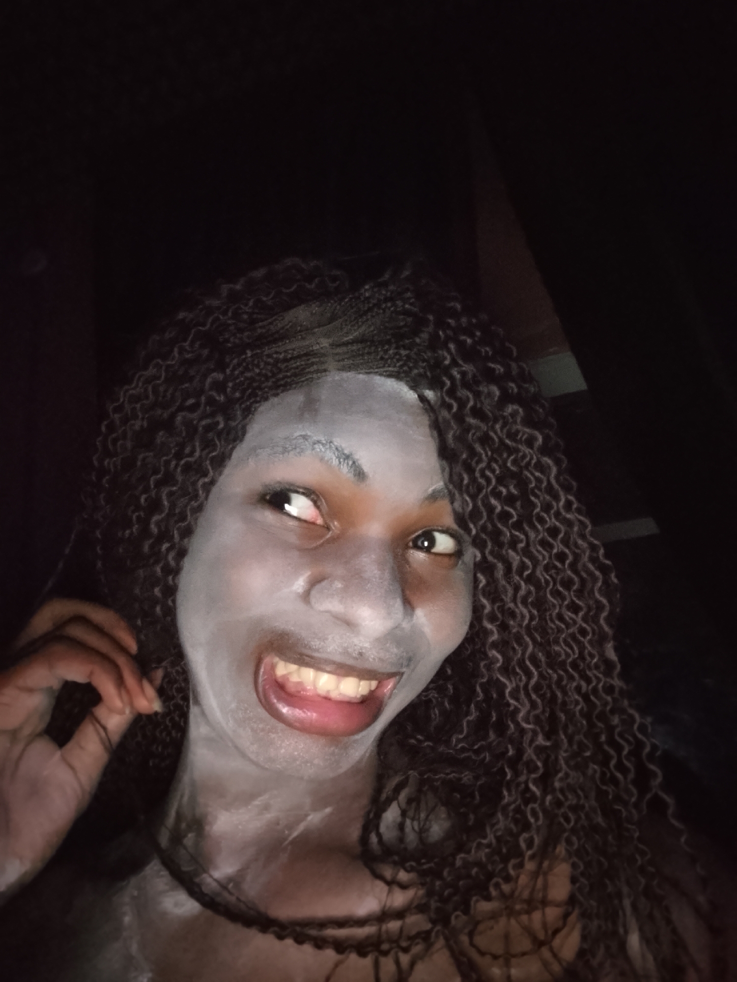 A close-up portrait features a person, likely a woman with dark skin, whose face and visible neck/shoulder area are covered in a light grey or white substance, resembling a facial mask or special effects makeup. She looks upwards and to the right with a wide, open-mouthed smile, revealing her teeth and tongue, and her eyes are bright, conveying a playful or excited expression. Her dark, curly braided hair frames her face, with strands falling down her shoulders. One hand, with dark-painted nails, is visible near the left side of her face, seemingly adjusting her hair or touching her jawline. The background is completely dark, indicating the image was taken at night or in a very dim setting. A direct light source illuminates her face, creating highlights on the applied substance and emphasizing her features. No text is visible in the image.