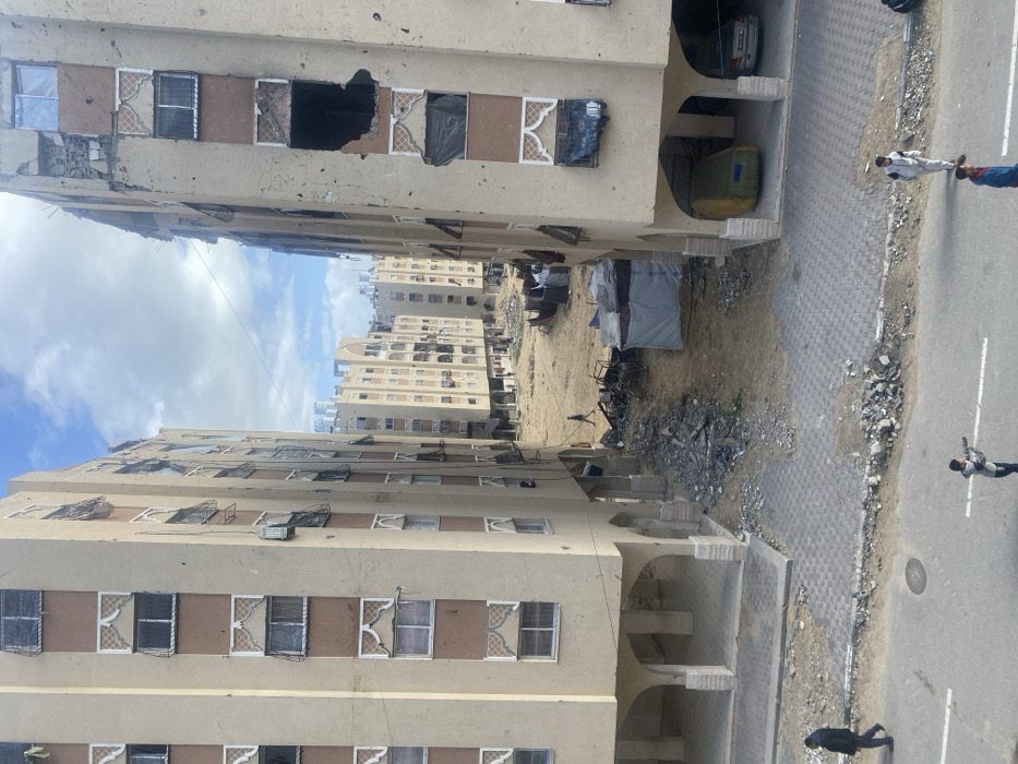 The image captures a residential area in Khan Yunis, Palestinian Territory, from an elevated perspective, depicting widespread damage. Two multi-story, light beige apartment buildings with brown vertical accents and ornate window designs flank a central unpaved area. The building on the right displays severe structural damage, with large sections of its facade and multiple windows destroyed, revealing darkened interiors. The building on the left shows extensive window damage, including broken glass and makeshift coverings.
Between these two primary structures, a wide expanse of dirt and sand is heavily littered with rubble, twisted metal, and debris. Dark, amorphous piles of what appears to be burnt material are present, along with a small, white, tent-like structure. A lone male figure in dark clothing walks across this debris-strewn area towards the background. In the distance, additional multi-story residential buildings, also exhibiting damage, line the horizon, with several white water tanks visible on their rooftops.
The foreground features a paved street with white markings. Adjacent to the street, a sidewalk of grey interlocking bricks is partially covered with debris and dirt. On the street and sidewalk, four individuals are visible: one male in dark attire walks left on the sidewalk, one male in light-colored clothing walks right on the paved road, and two younger males stand on the road, one holding a stick or pipe. Underneath the ground floor arches of the right building, a partially covered, light green vehicle is parked next to a silver sedan. The sky is partly cloudy.